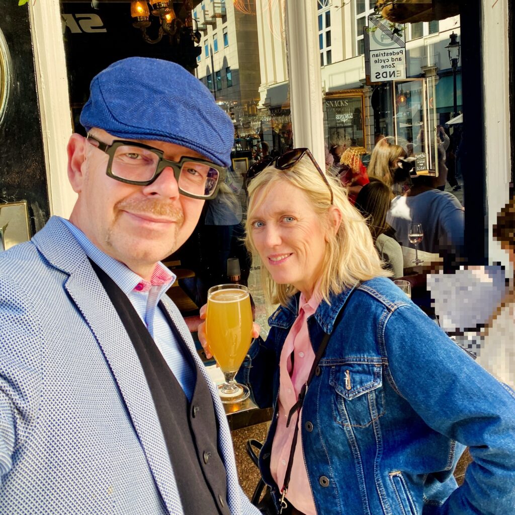 Man in a blue cap and blazer posing with a woman in a denim jacket holding a drink outside a café.