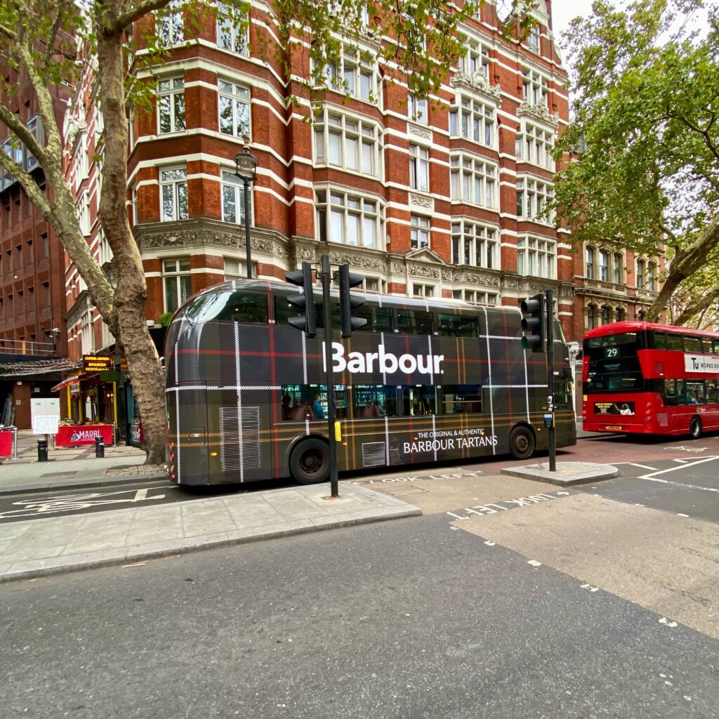 A double-decker bus wrapped in a Barbour tartan design parked at a traffic light in an urban setting.