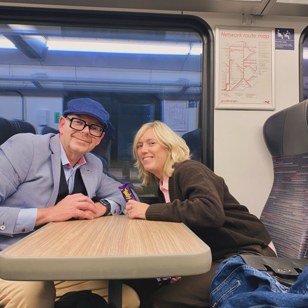 Two individuals seated at a table on a train, one wearing glasses and a cap, the other holding a chocolate bar.