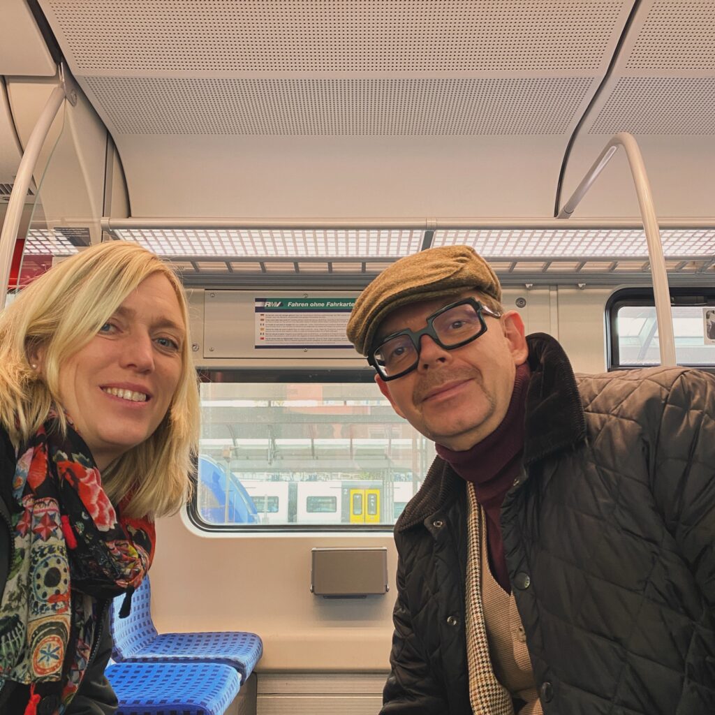 Two individuals seated on a train, one wearing a flat cap and glasses, the other with long blonde hair and a patterned scarf.