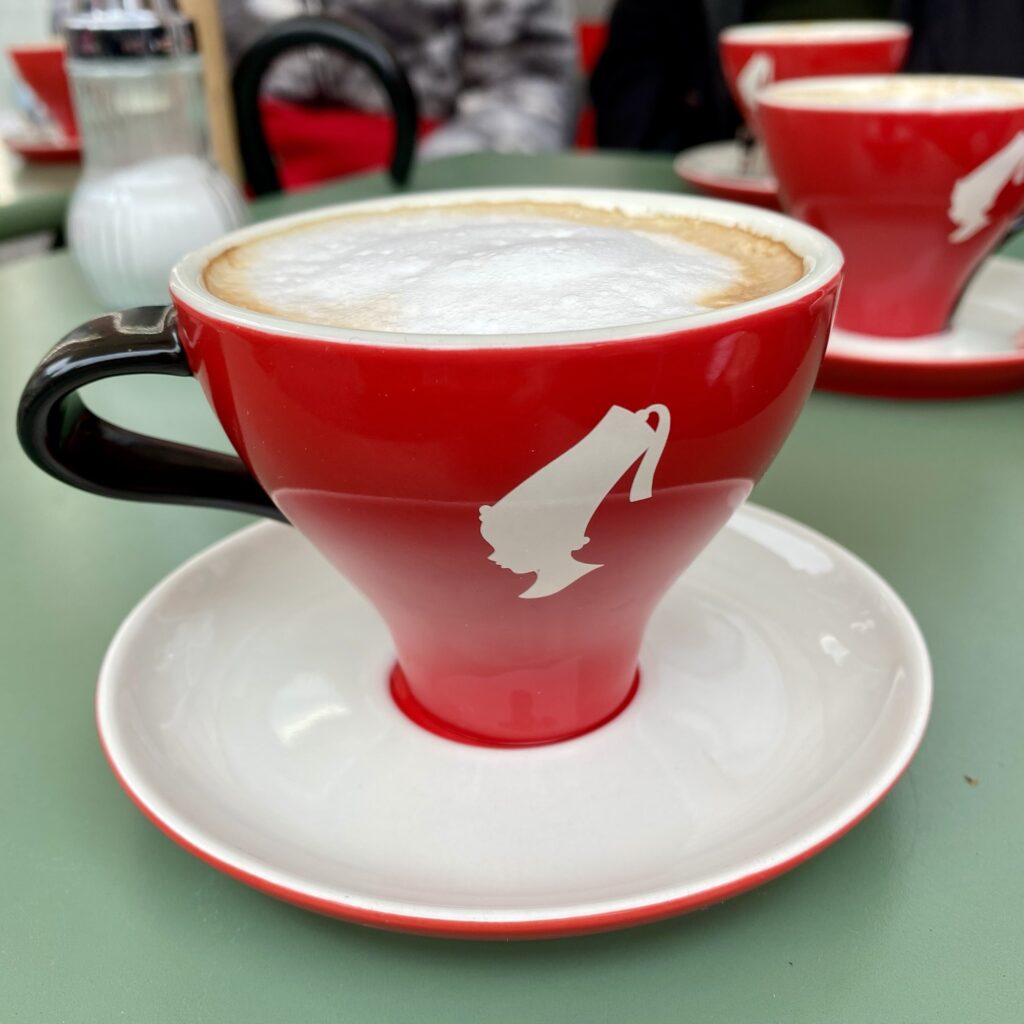 Red coffee cup with a white logo on a saucer, filled with cappuccino topped with frothy milk.