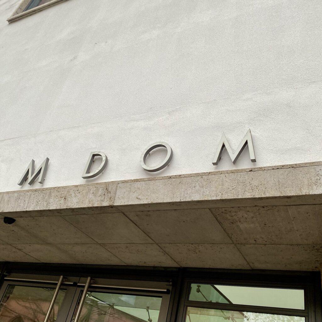 Signage displaying the letters 'M DOM' mounted on a textured wall above an entrance.