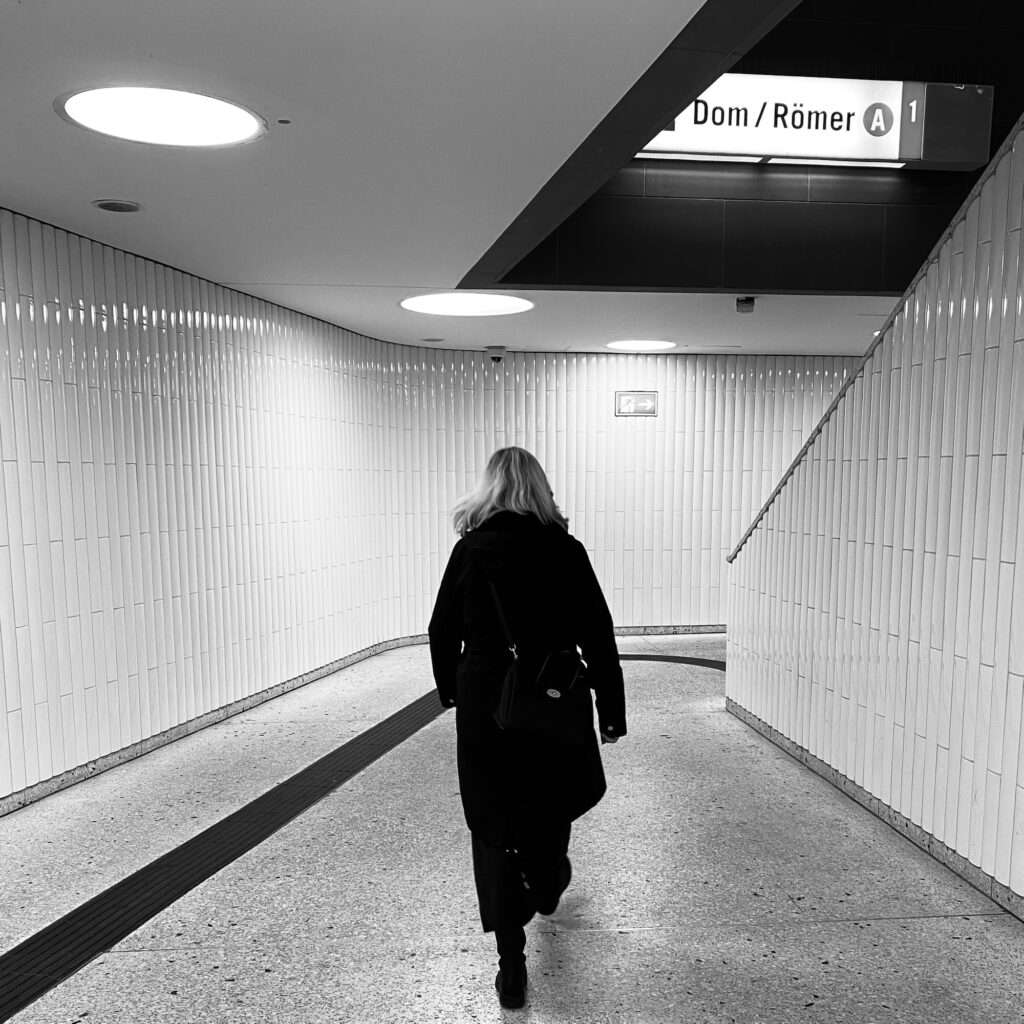 A woman walking through a subway station corridor with white tiled walls and overhead lighting.