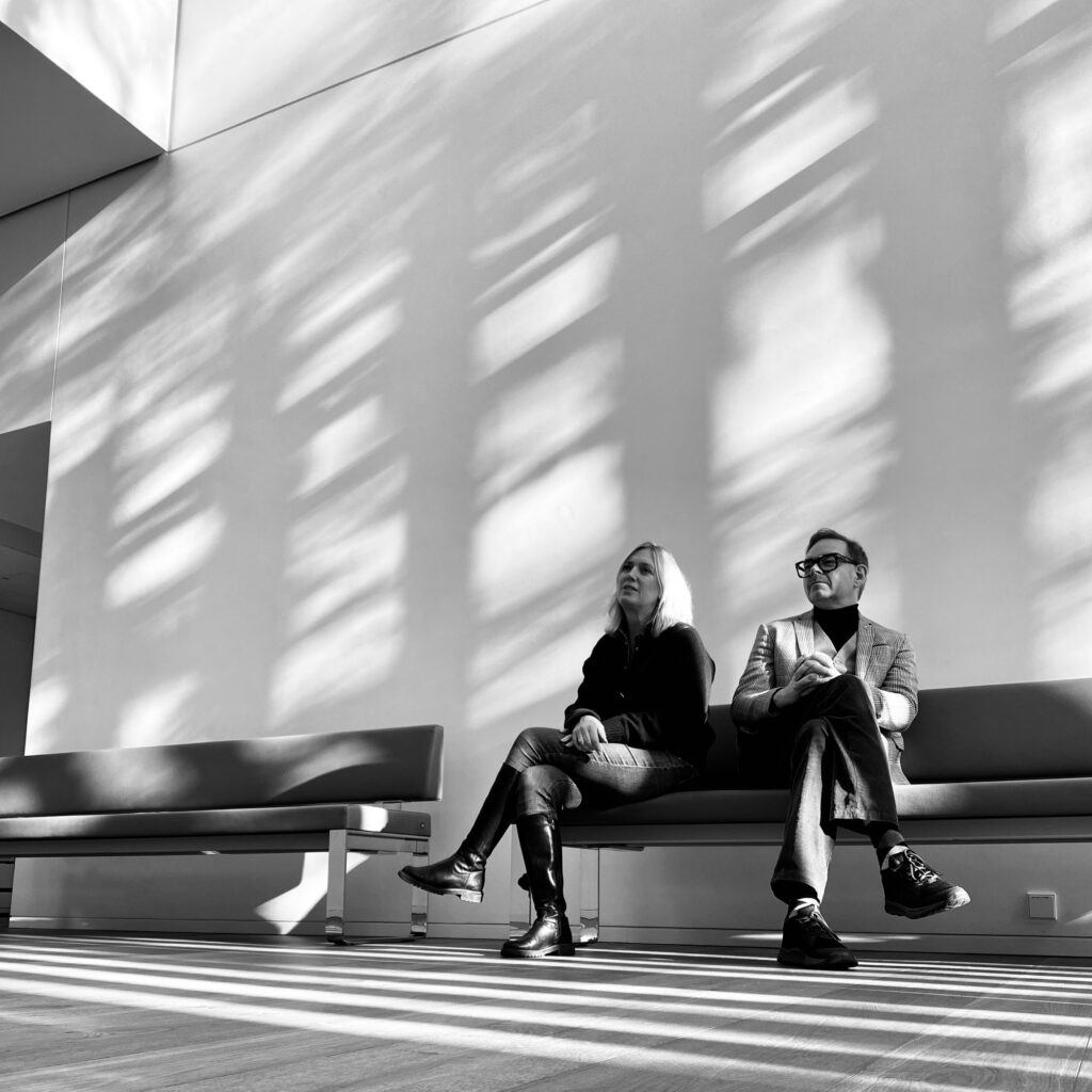 Two individuals seated on a bench in a brightly lit space with shadows cast on the wall.
