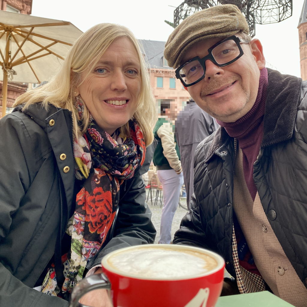 A man and a woman smiling at the camera with a coffee cup in front of them at an outdoor café.