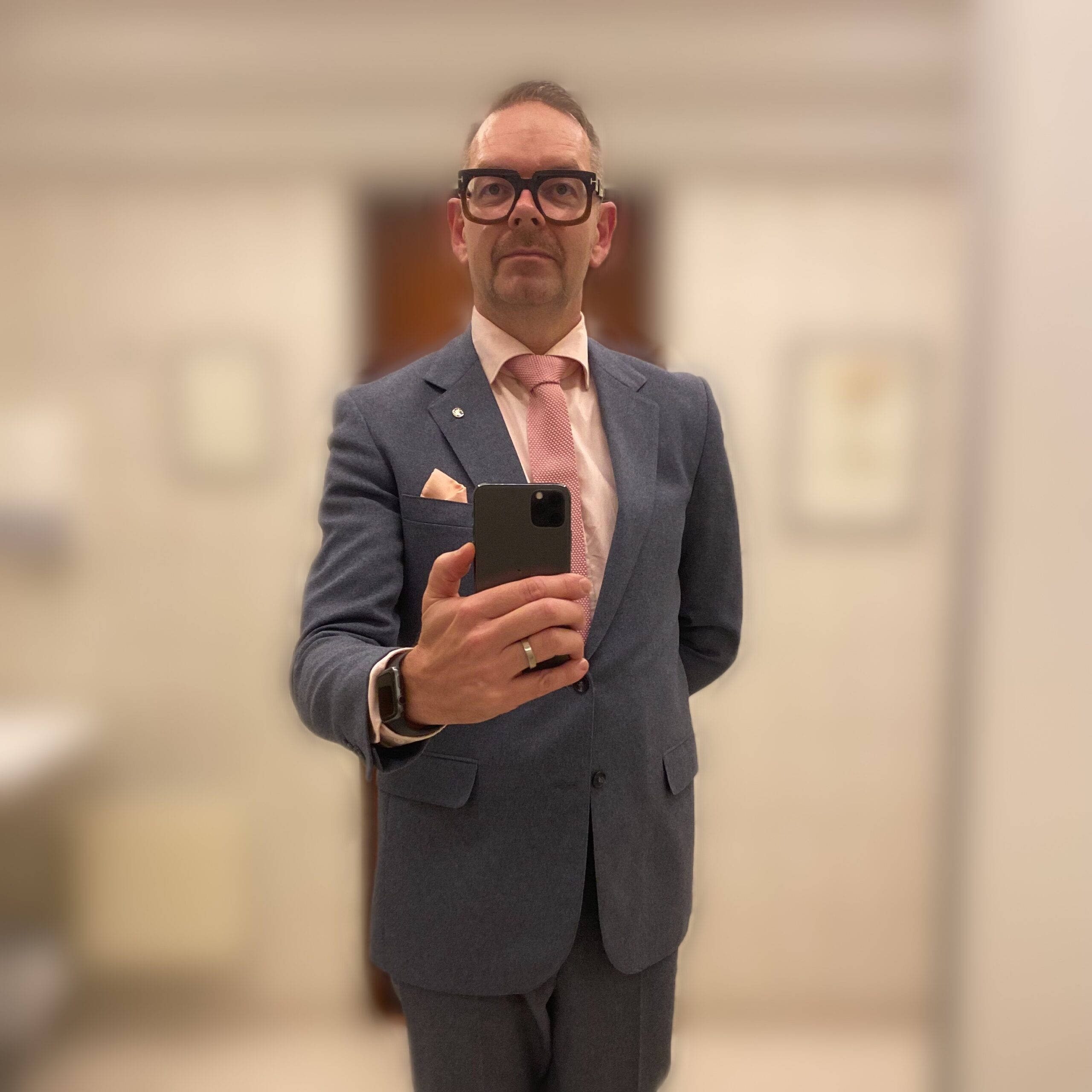 Man in a tailored suit holding a smartphone, standing in a mirror reflection.
