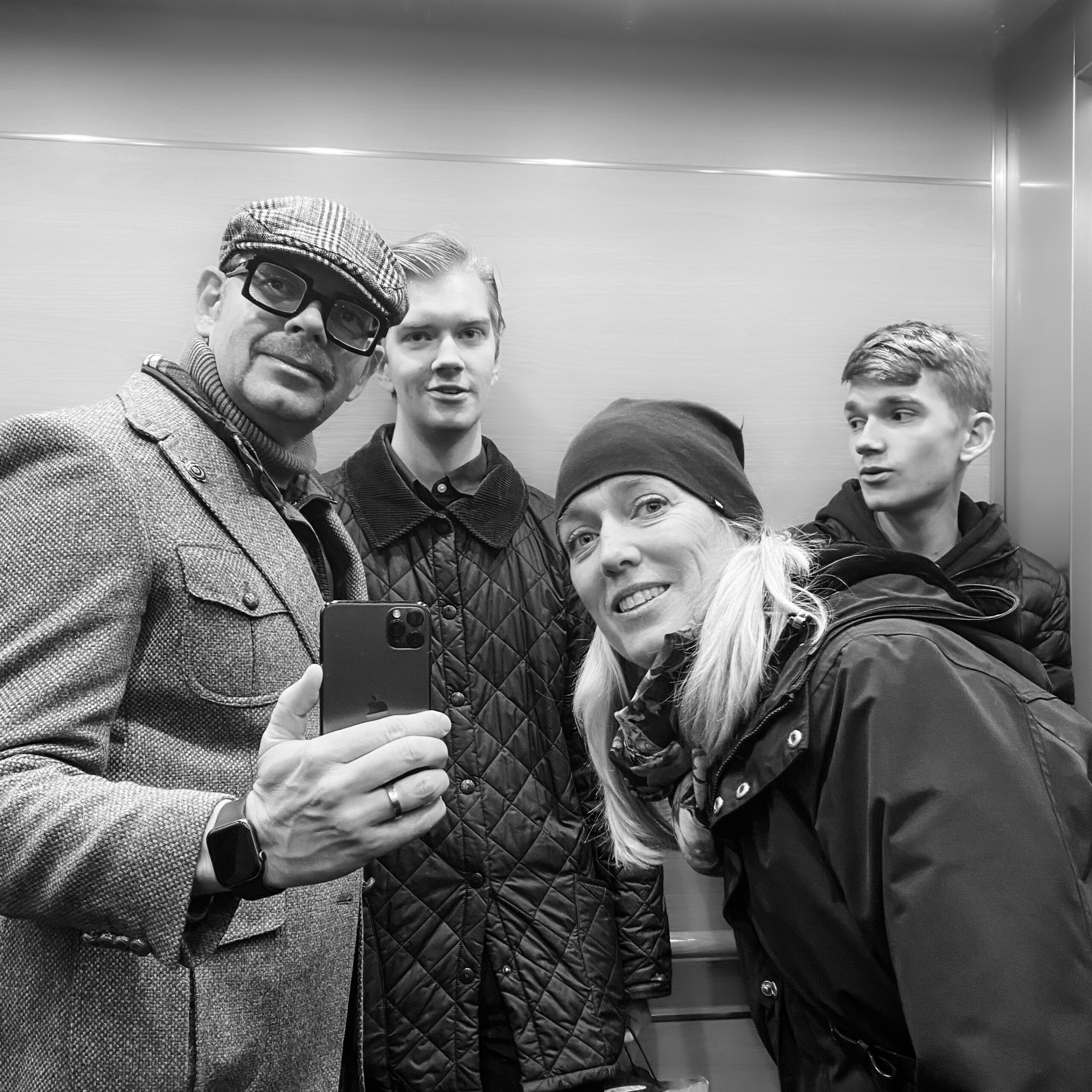 Group of four people taking a selfie in a lift, dressed in winter attire with a smartphone.