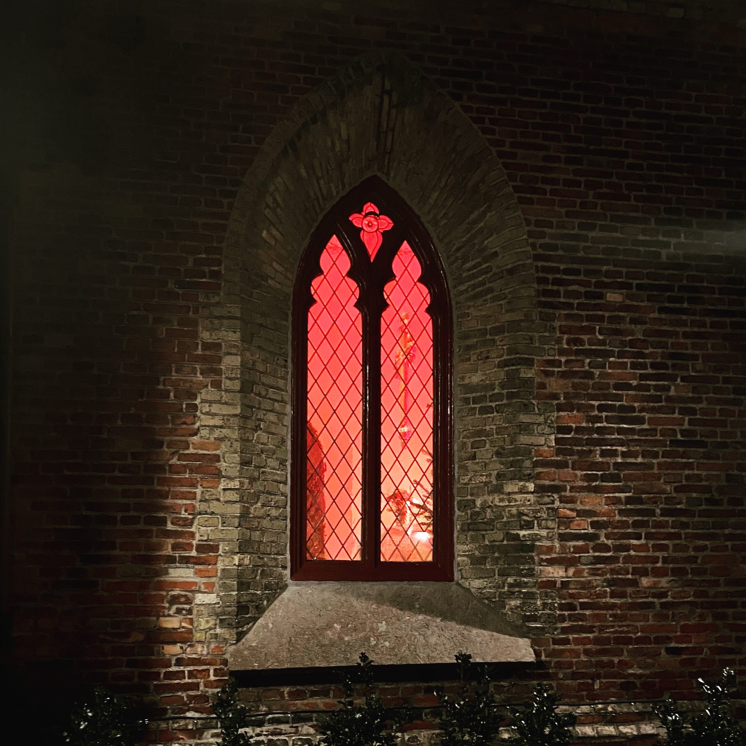 Gothic-style window with red stained glass in a brick wall, illuminated from within.