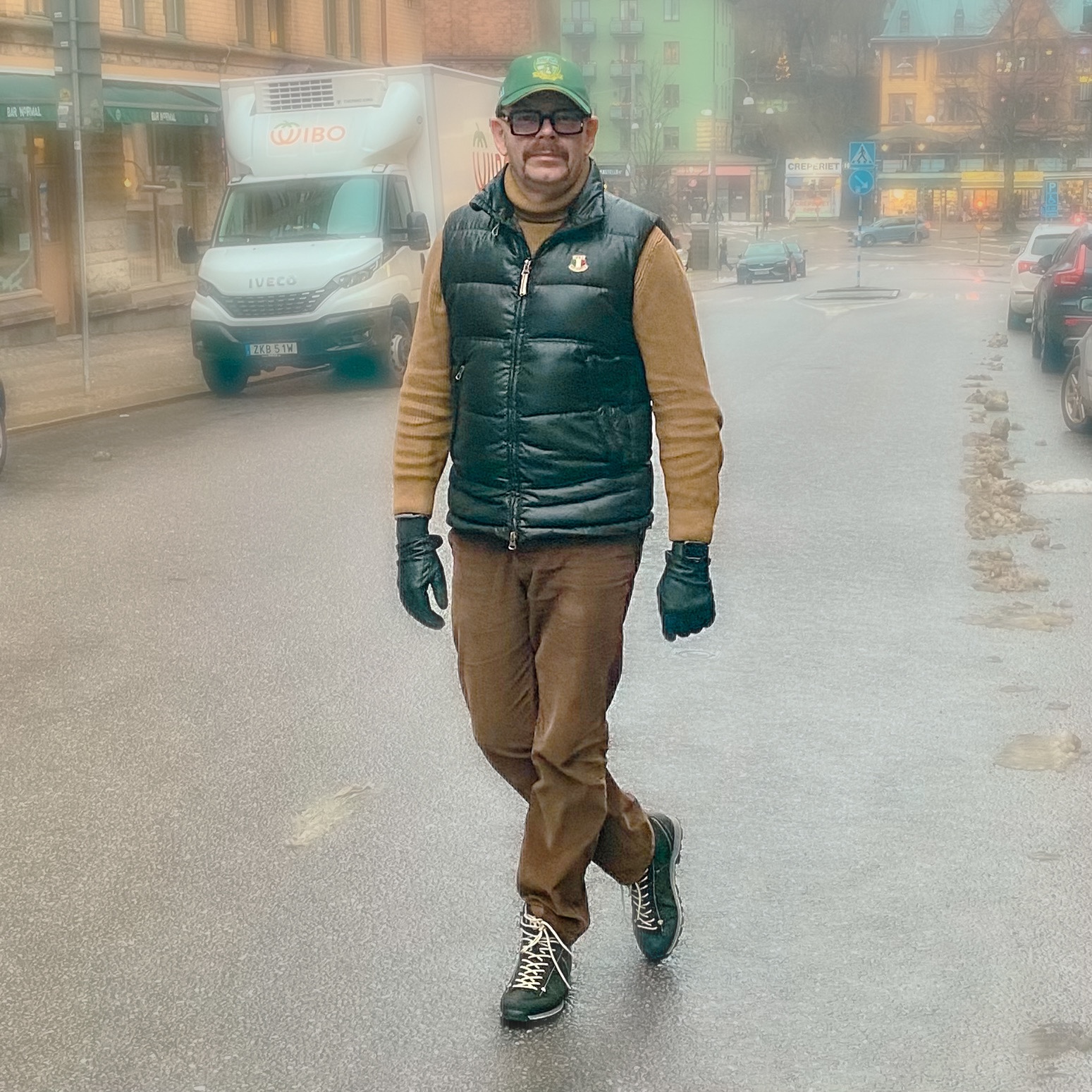 Man walking on a street wearing a black vest, brown sweater, and gloves in a foggy urban environment.