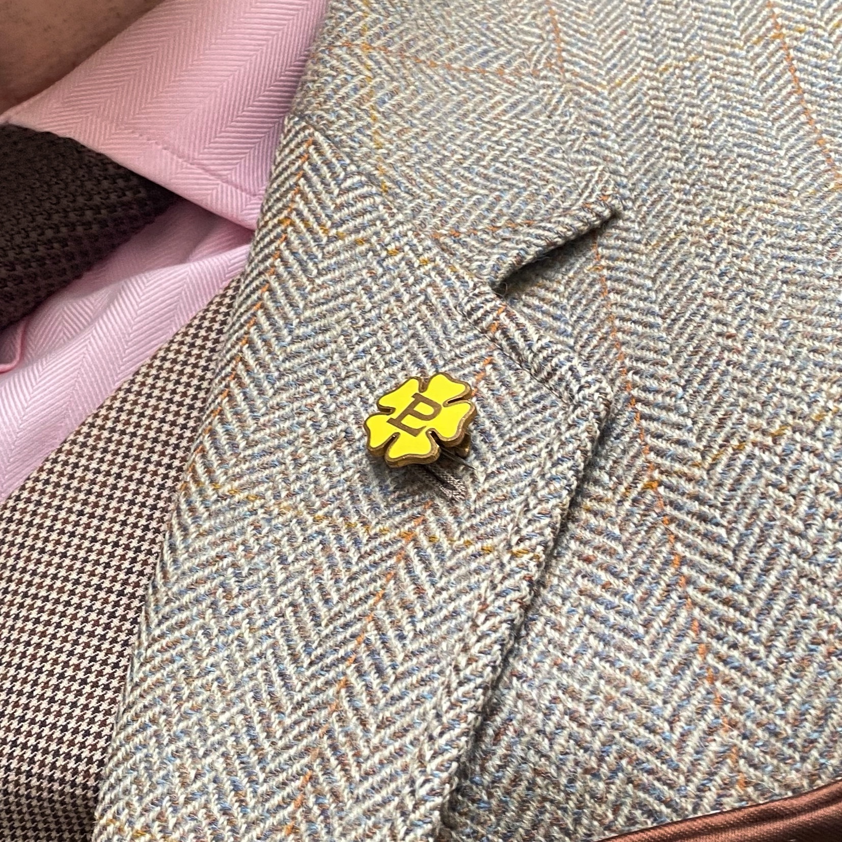 Yellow enamel pin shaped like a primrose attached to a grey herringbone jacket lapel.