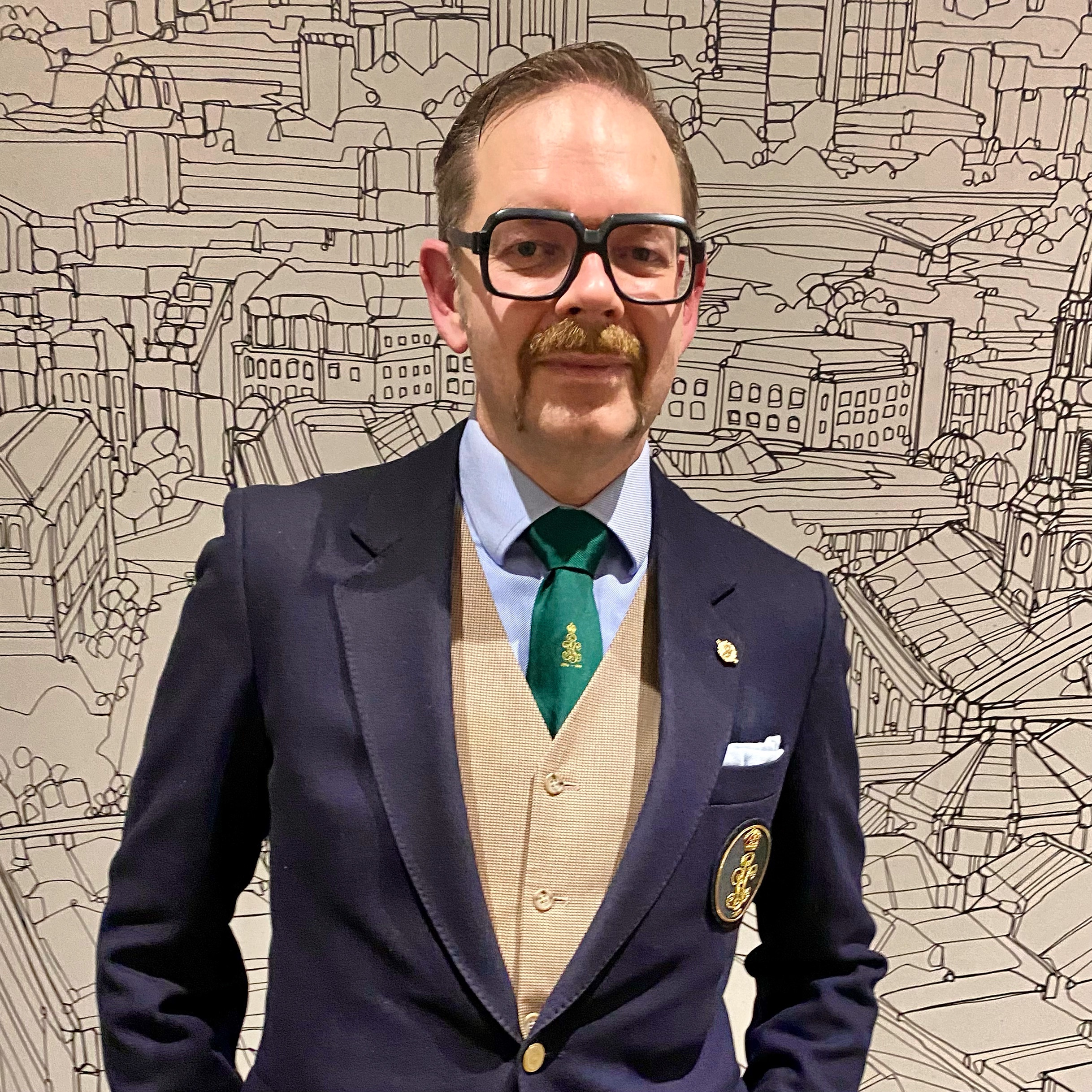 Man wearing a blazer and waistcoat with a green tie, standing in front of a detailed cityscape mural.