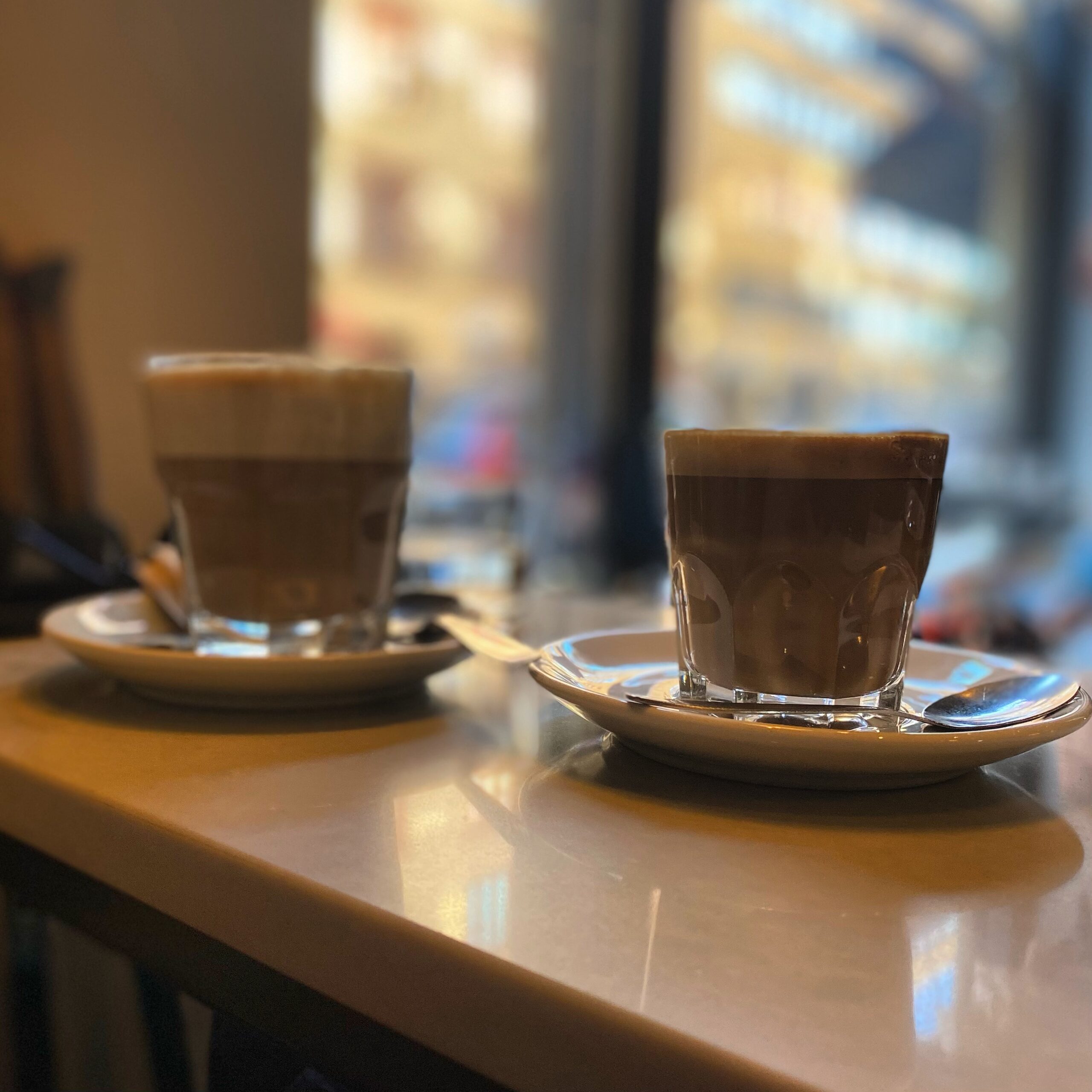 Two glasses of coffee on saucers, one with frothy milk and the other dark, placed on a table.