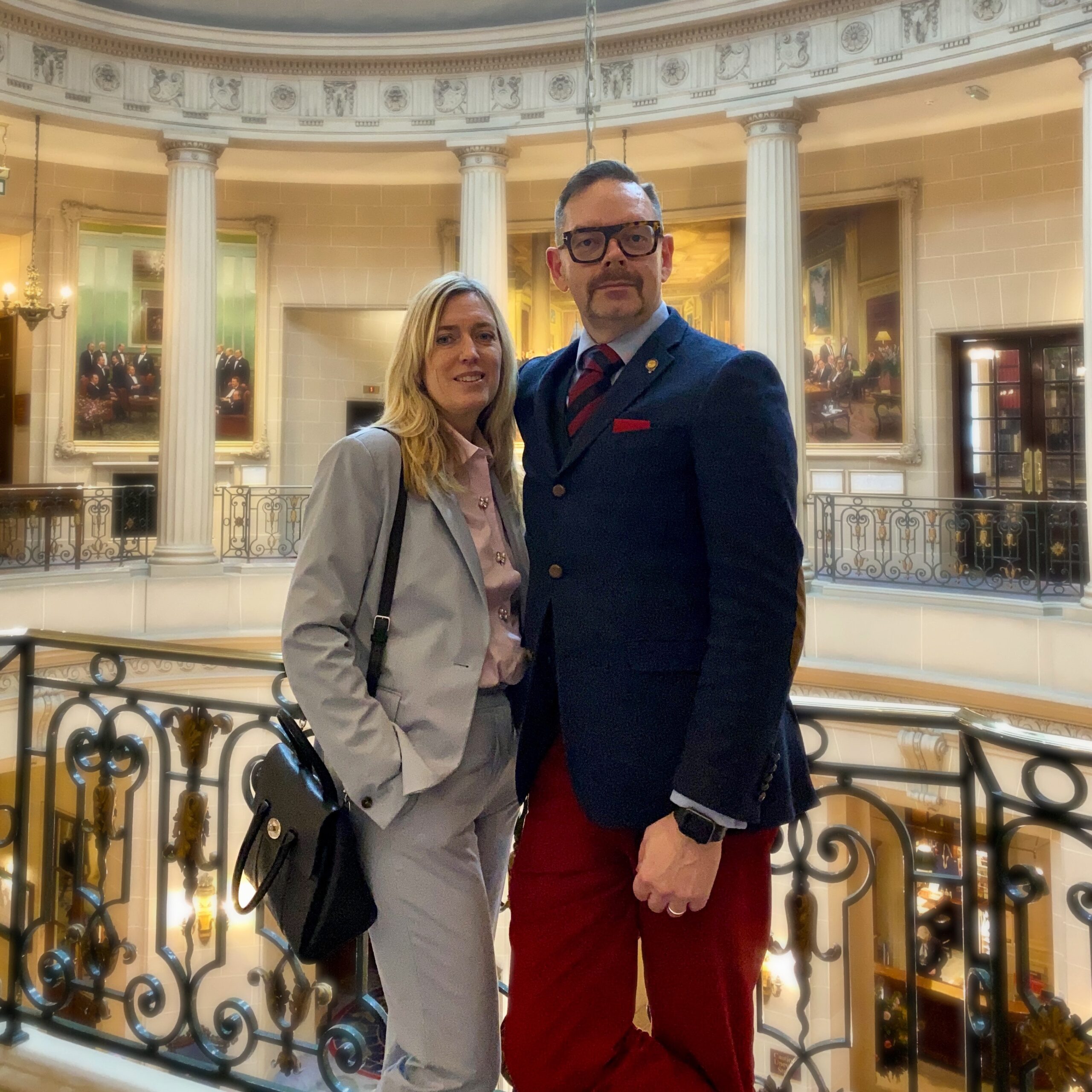 A man in a suit with red trousers and a woman in a grey suit standing together in an elegant interior space.