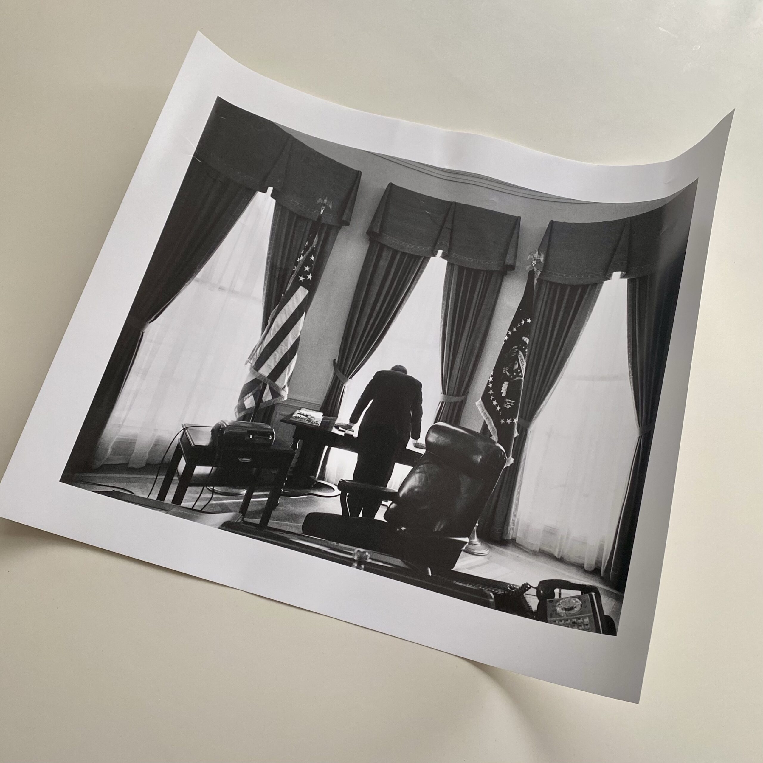 Black and white photograph of a man standing at a desk in an office, with curtains and an American flag visible.