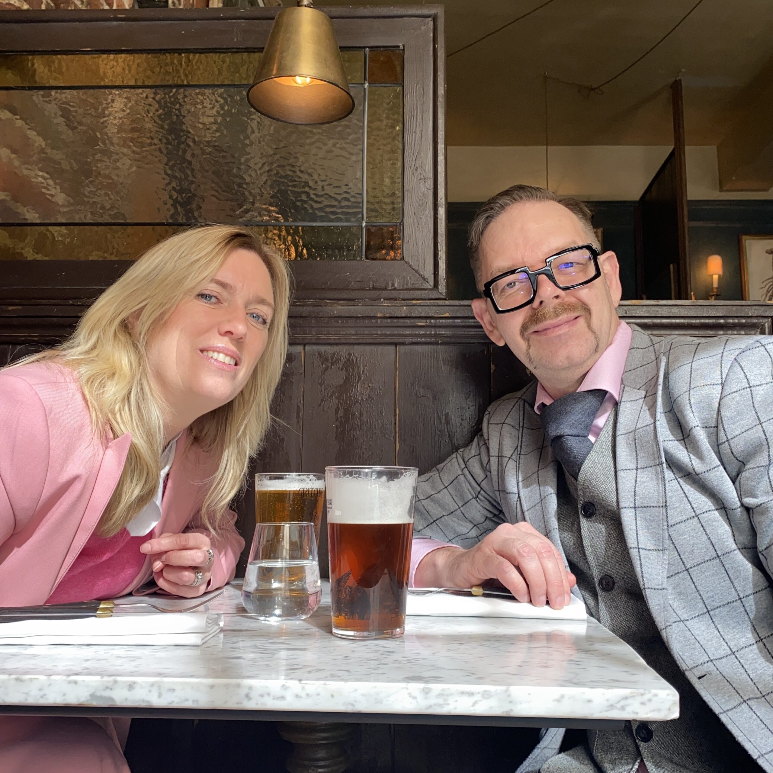 A man with glasses and a moustache and a woman in a pink suit sitting at a table with drinks in a restaurant.