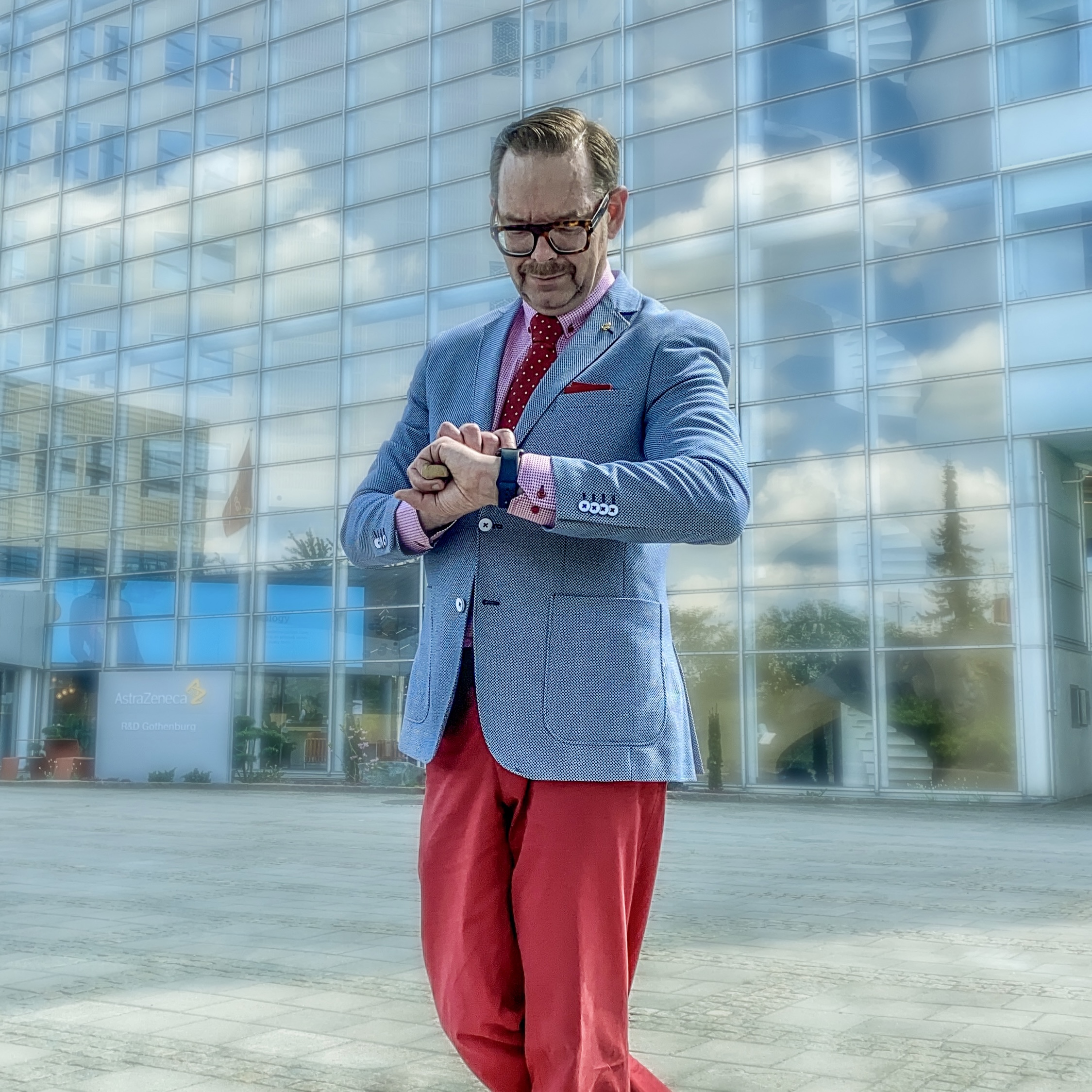 Man in a blue blazer and red trousers checking his watch outside a modern glass building.