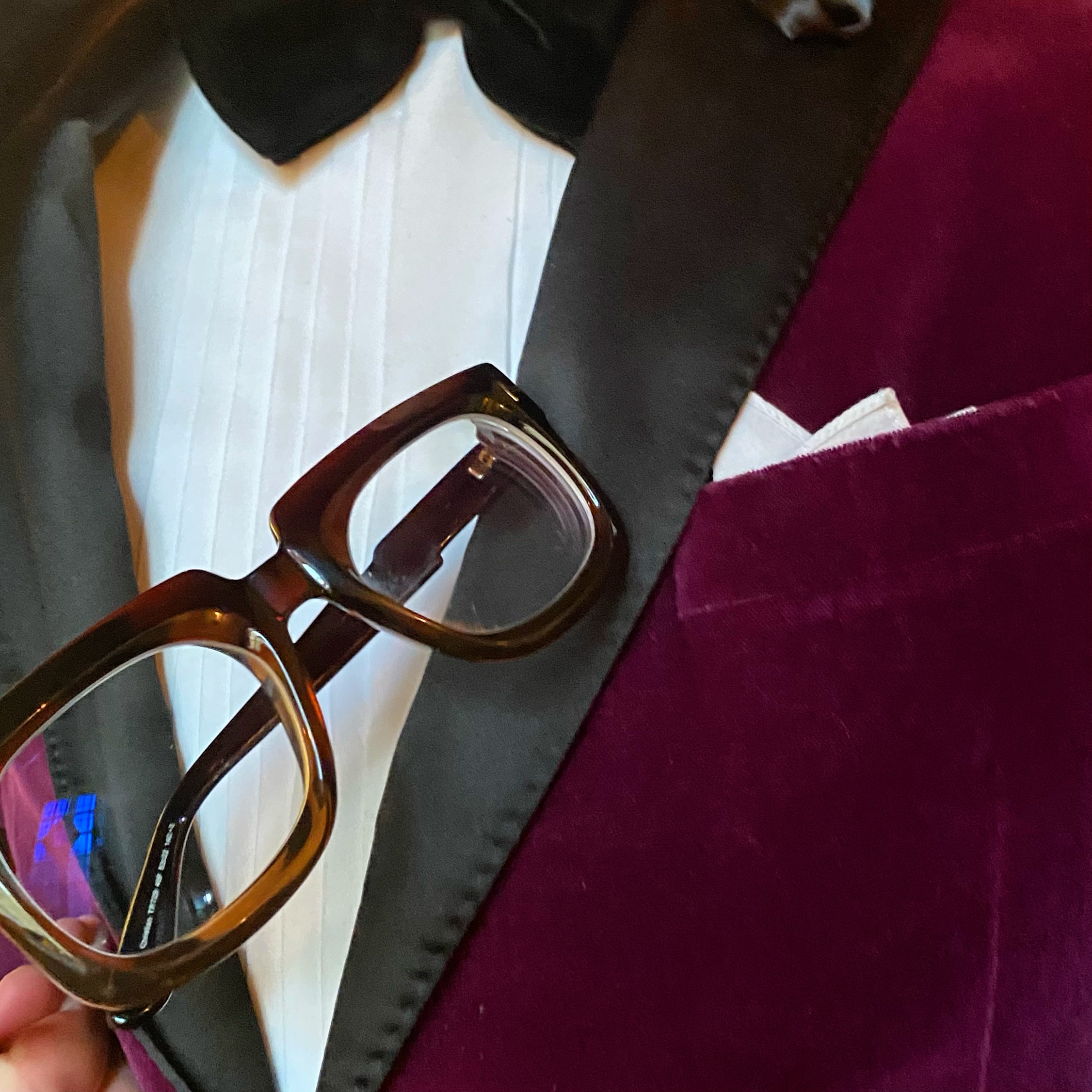 Close-up of a person holding stylish glasses next to a formal outfit with a bow tie and pocket square.