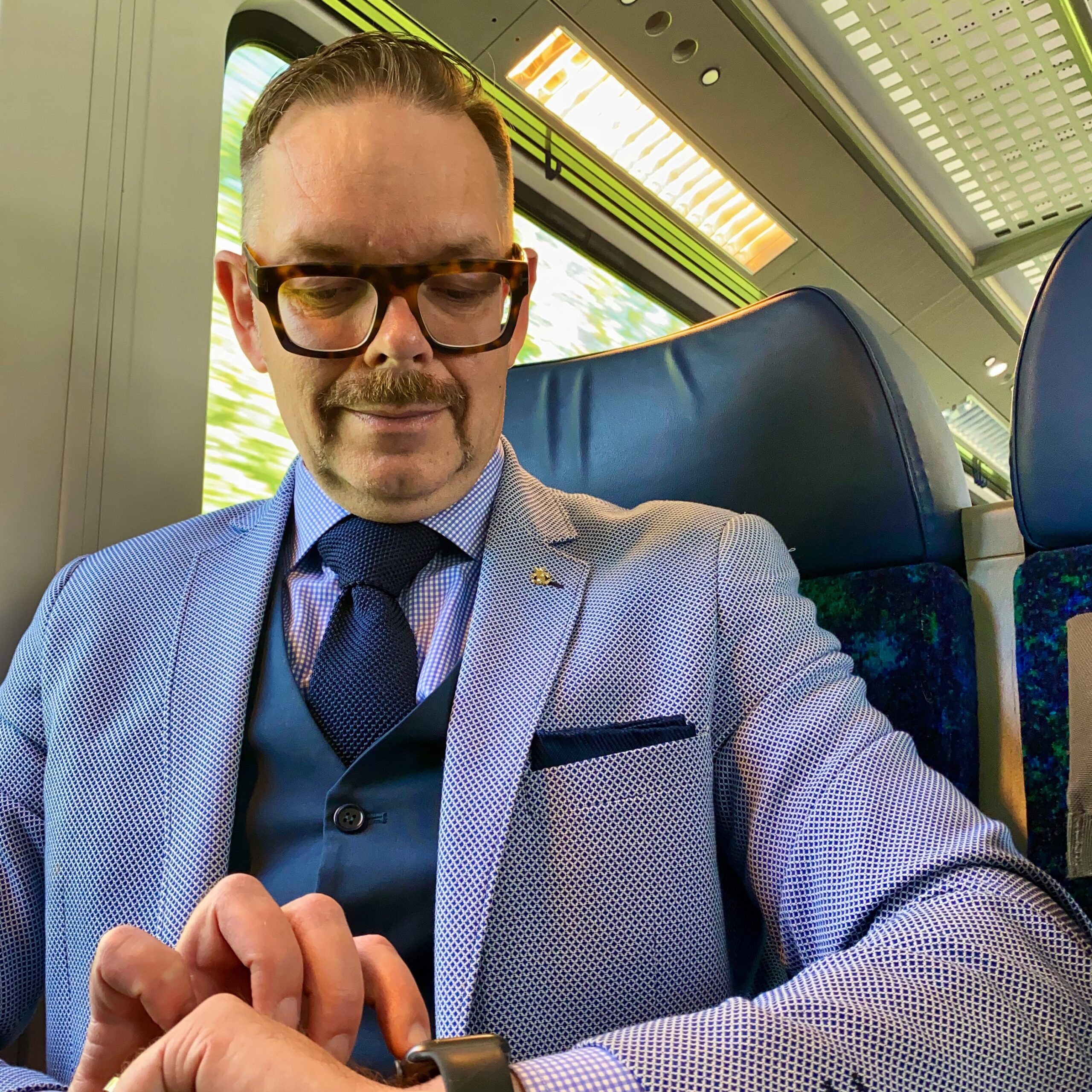 Man in a light blue suit checking his watch while seated on a train.