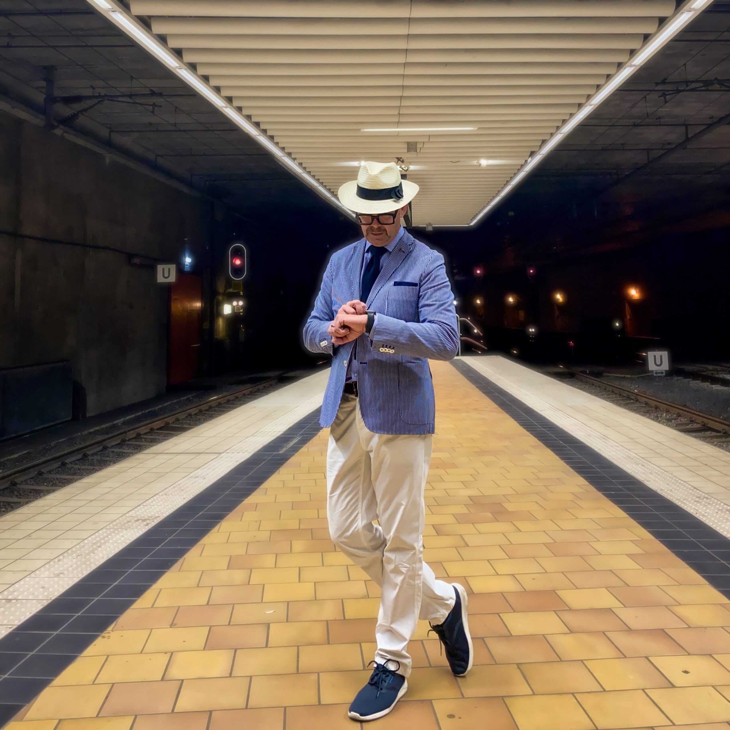 Man in a light blue blazer and hat checking his watch at a train station platform.