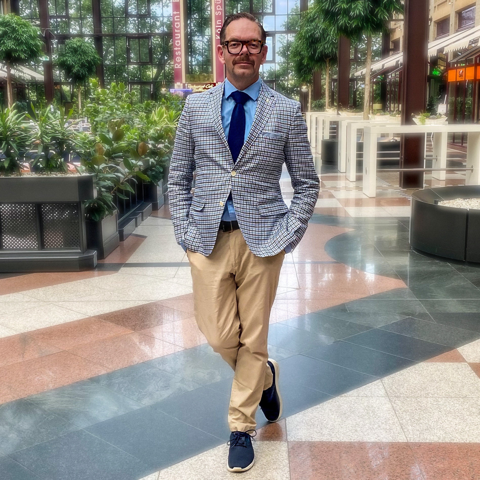 Man in a checked blazer and blue tie walking confidently in a modern indoor space with greenery.