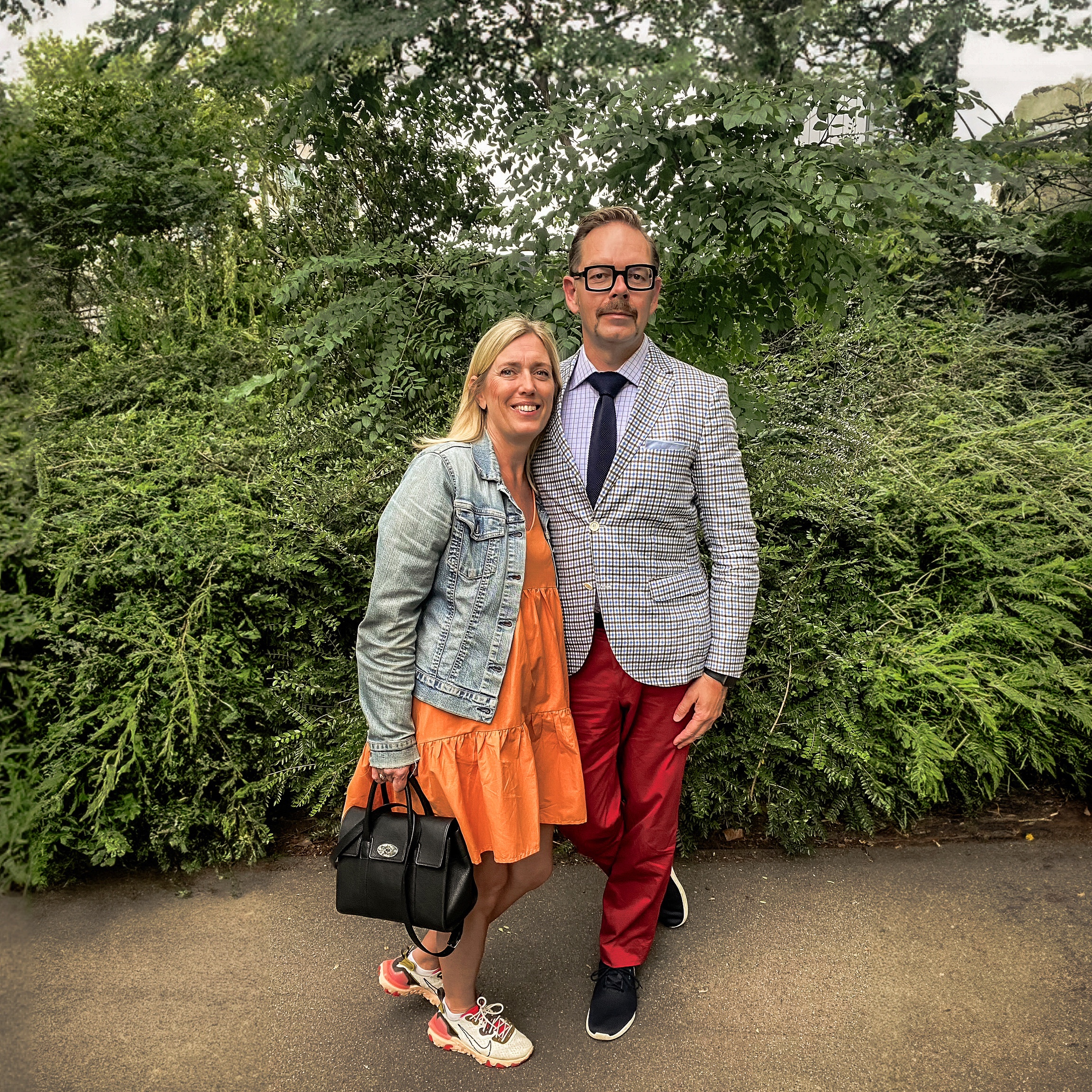A man in a checked blazer and red trousers stands next to a woman in a denim jacket and orange dress, both smiling.