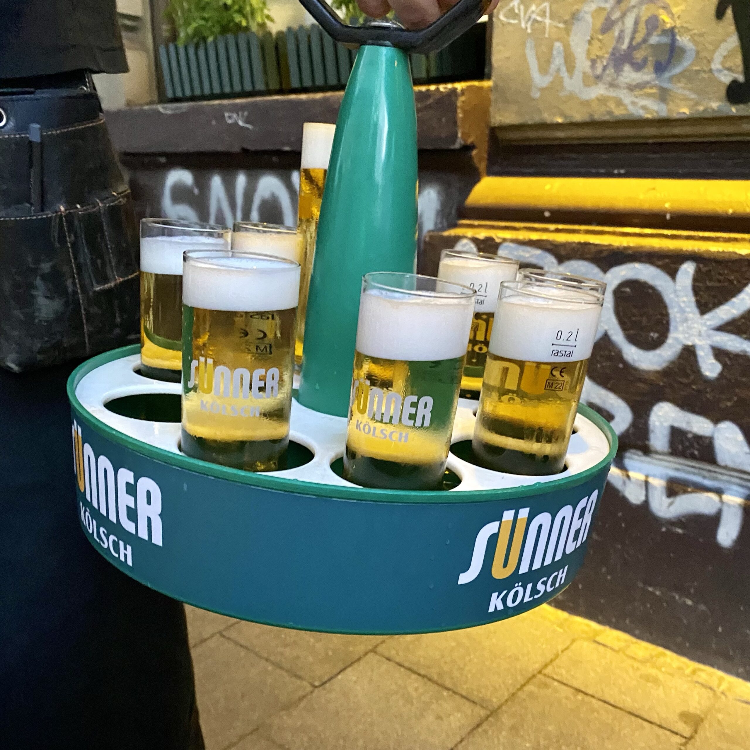 Tray holding multiple glasses of Kölsch beer, with a distinctive green handle and branding.