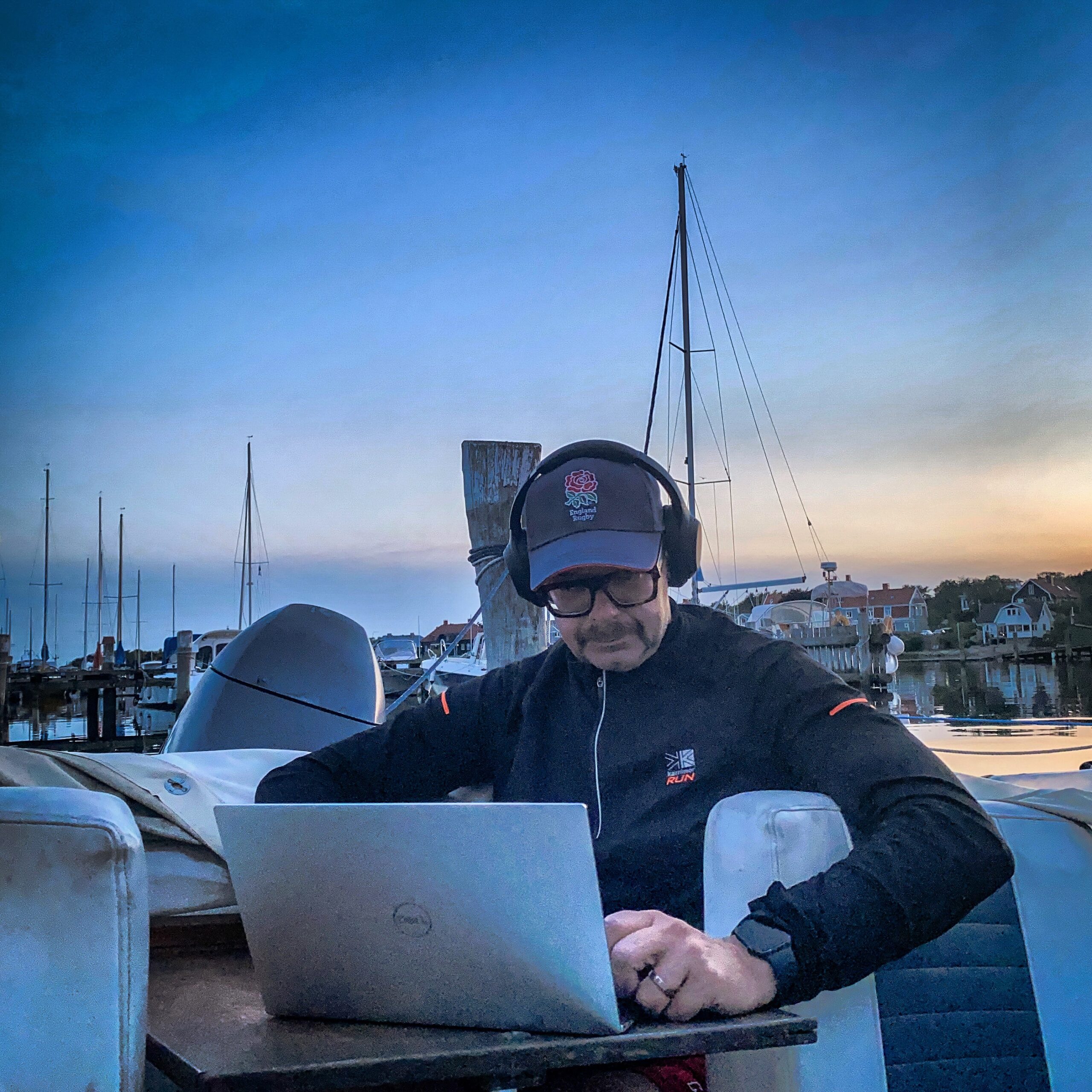 Man working on a laptop while wearing headphones, seated on a boat with masts in the background.