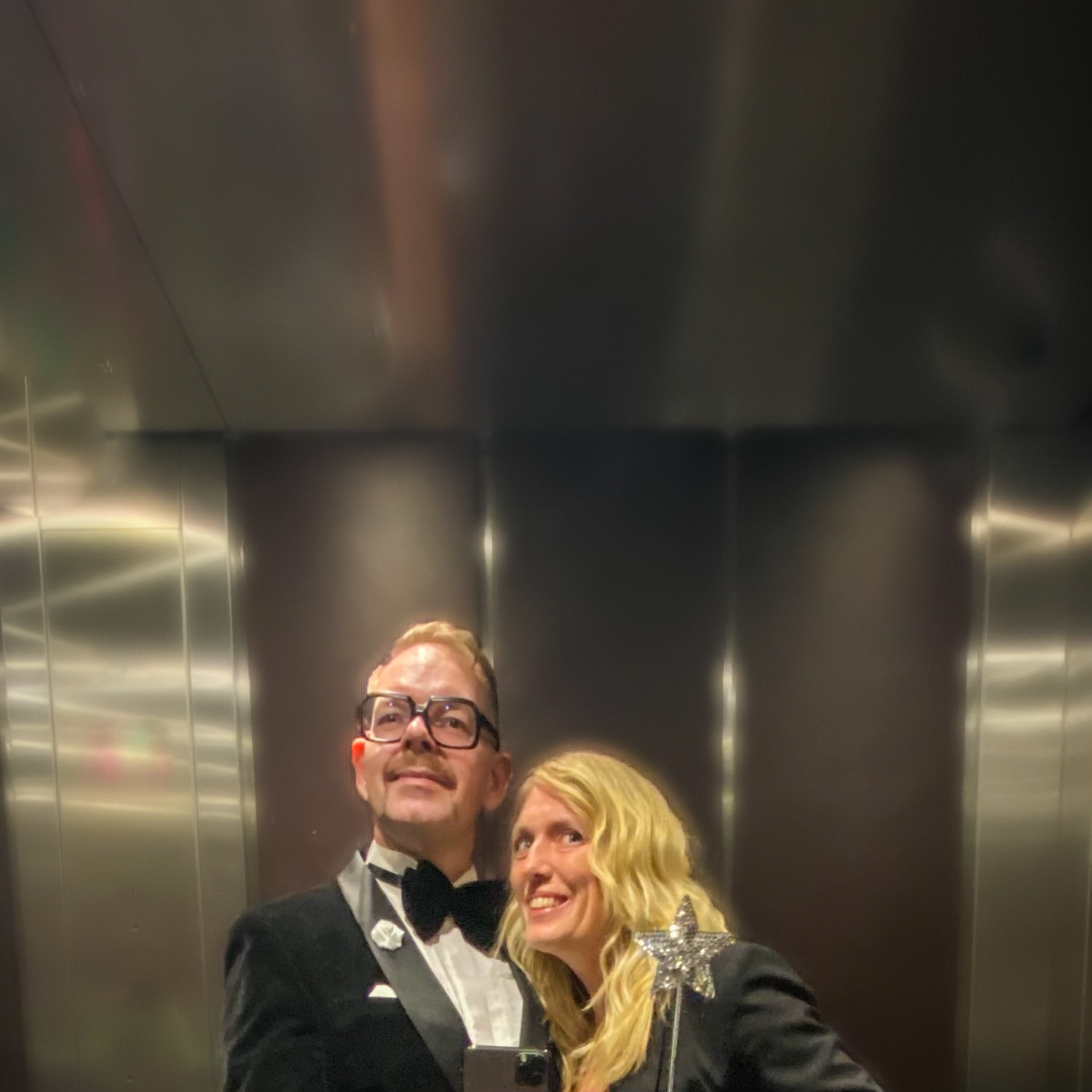 A man in a tuxedo and a woman in a black outfit posing together in a mirrored elevator.