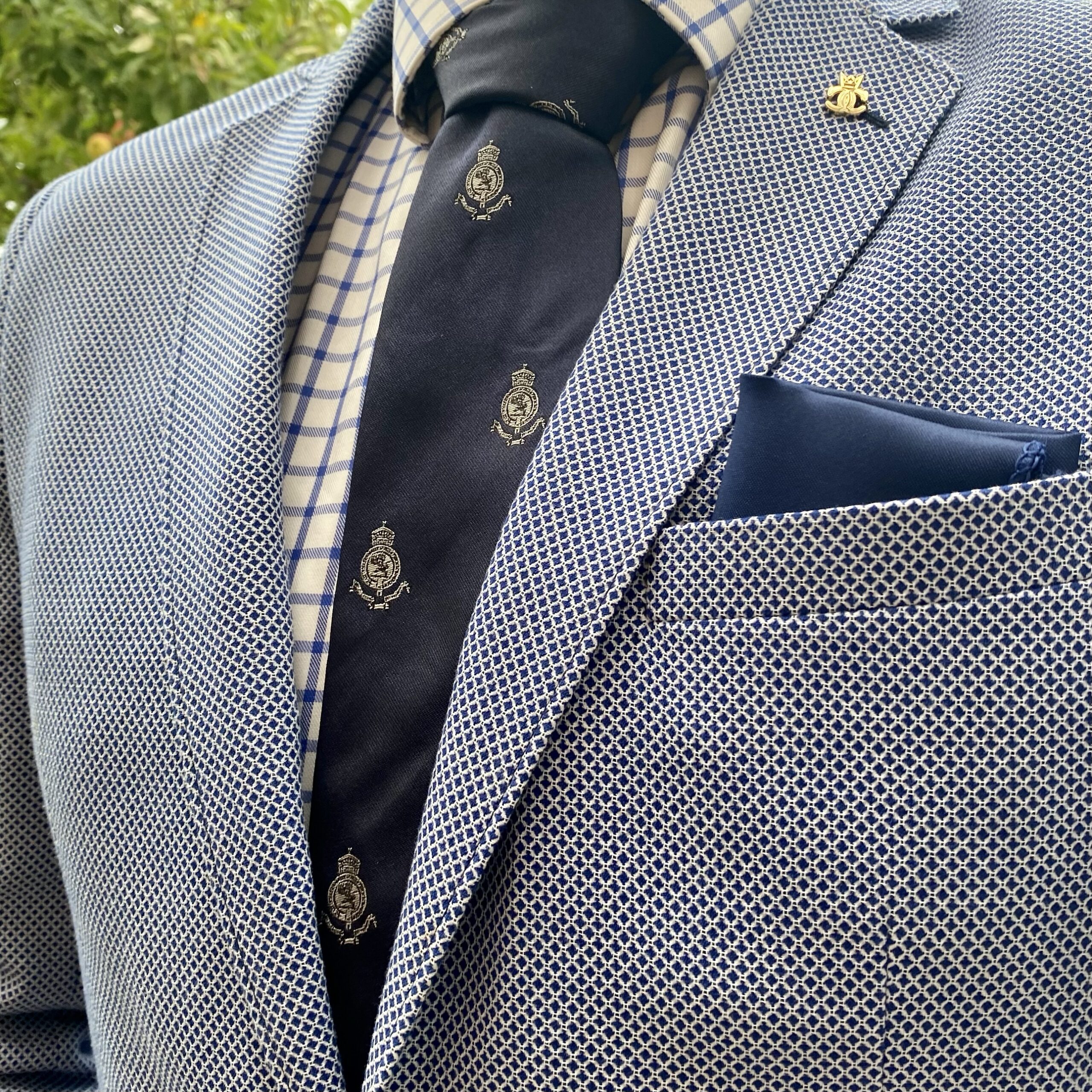 Close-up of a man in a patterned suit with a blue tie featuring a crest design and a pocket square.
