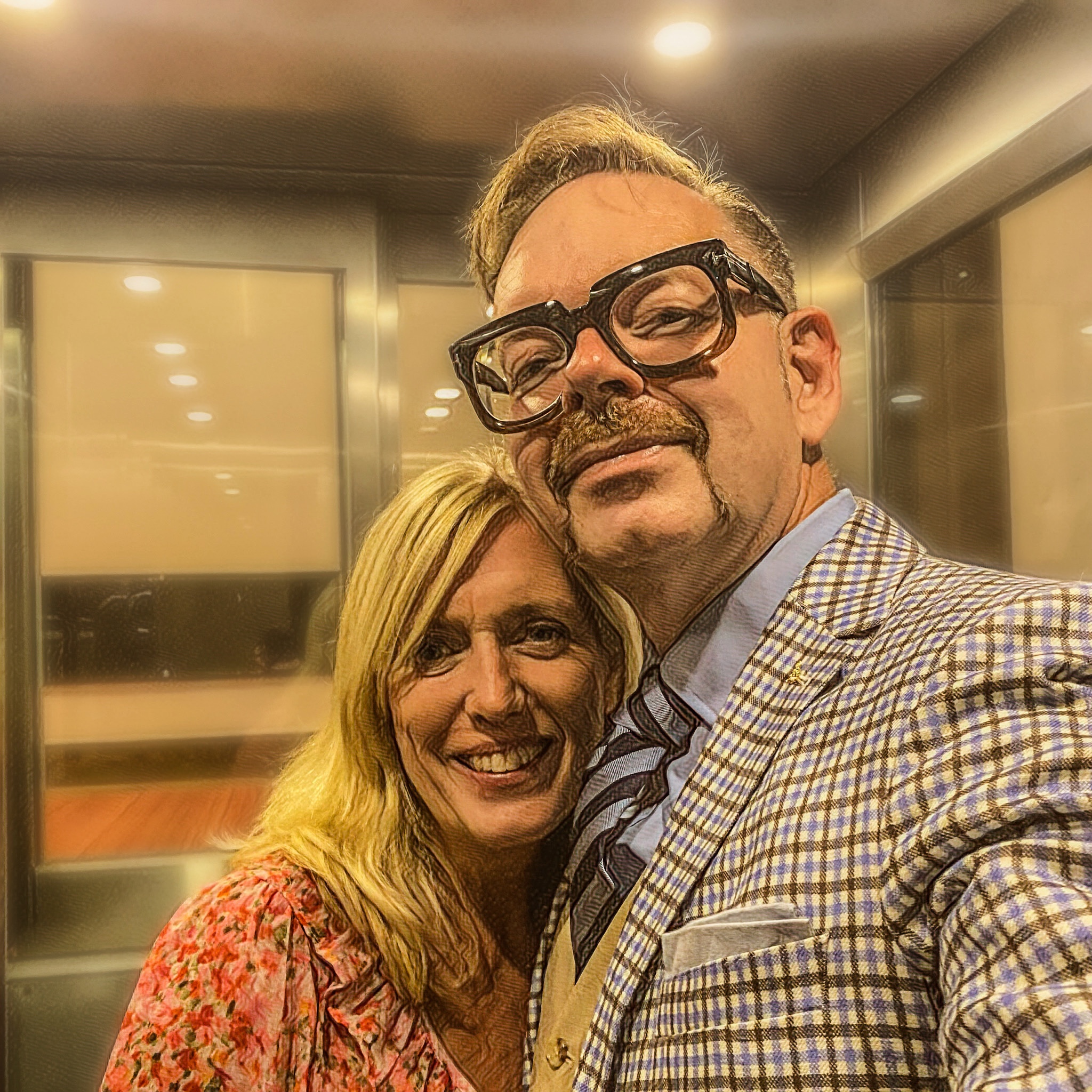 Man with glasses and a patterned suit standing next to a woman with long blonde hair in an elevator.