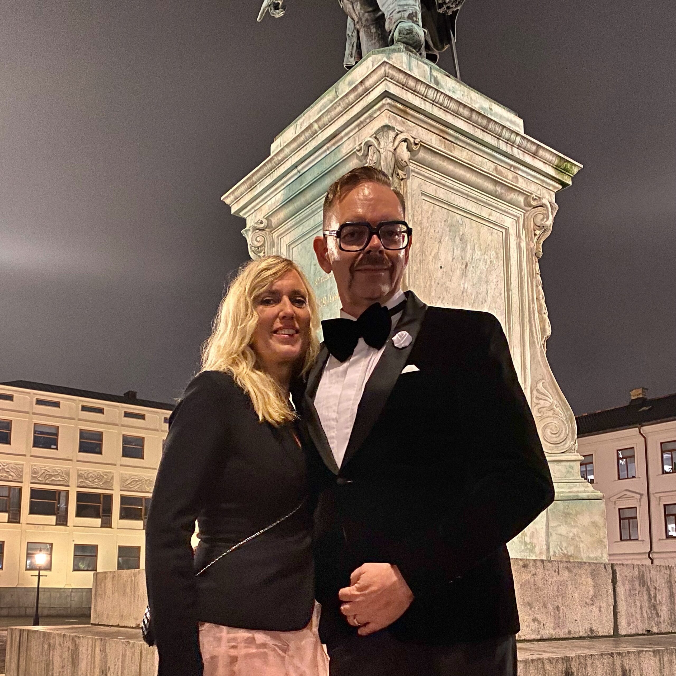 A man in a tuxedo and a woman in formal attire standing together in front of a statue at night.