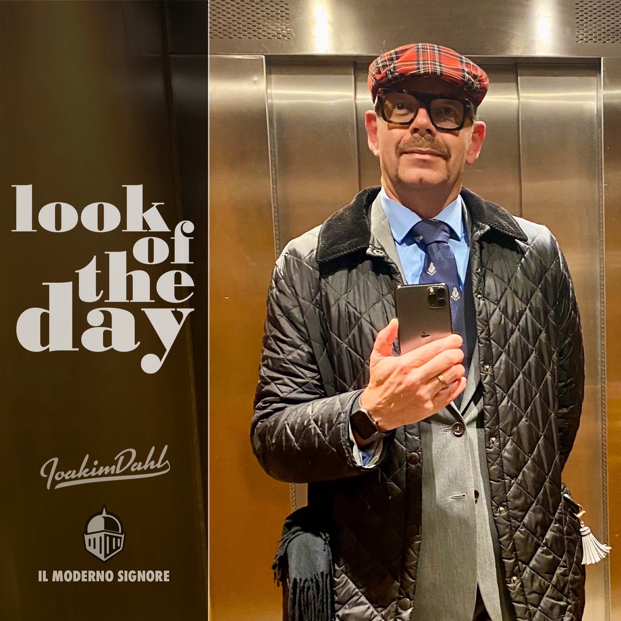 Man in a quilted jacket and flat cap holding a smartphone in an elevator mirror selfie.