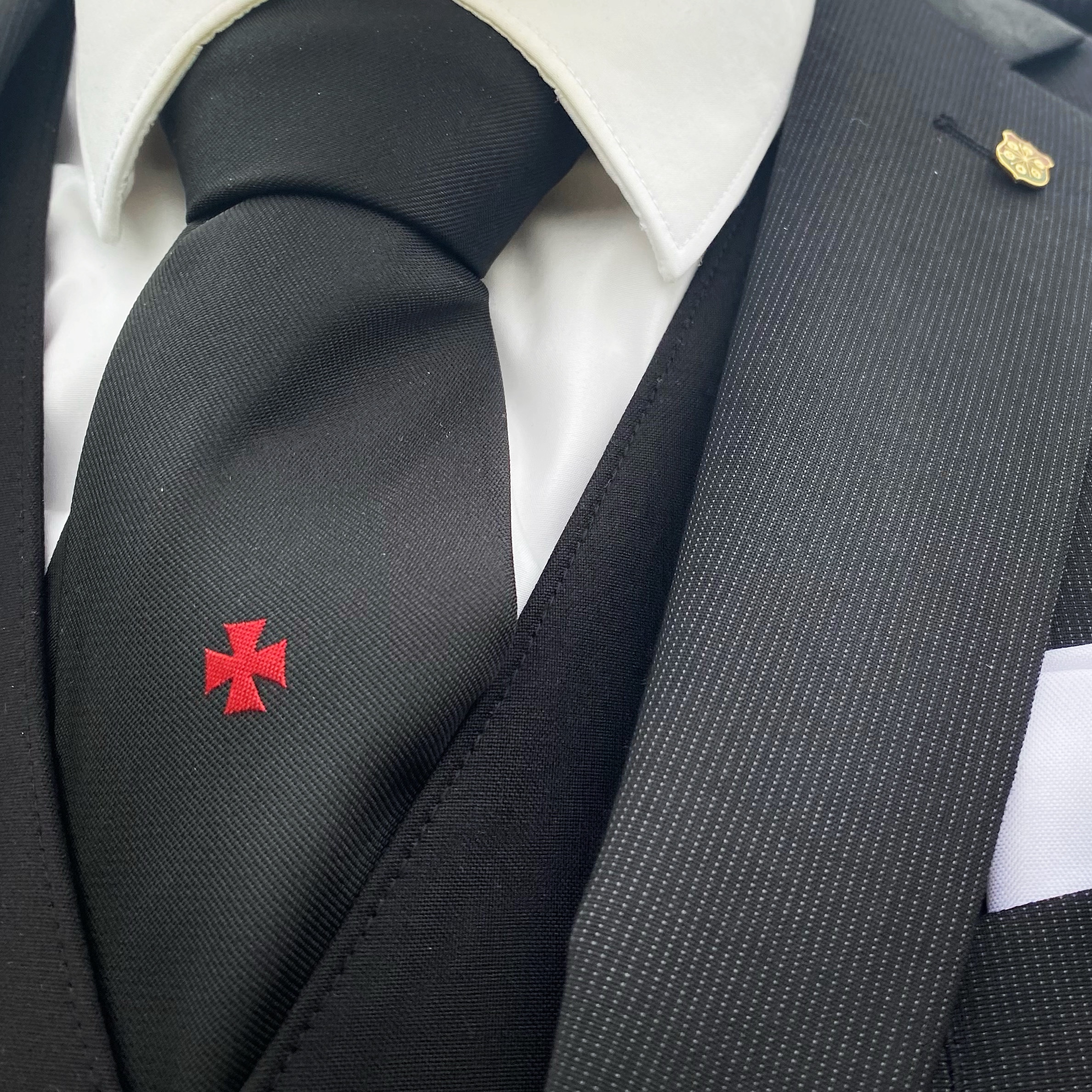Close-up of a black tie with a red cross emblem, worn with a formal suit and white shirt.
