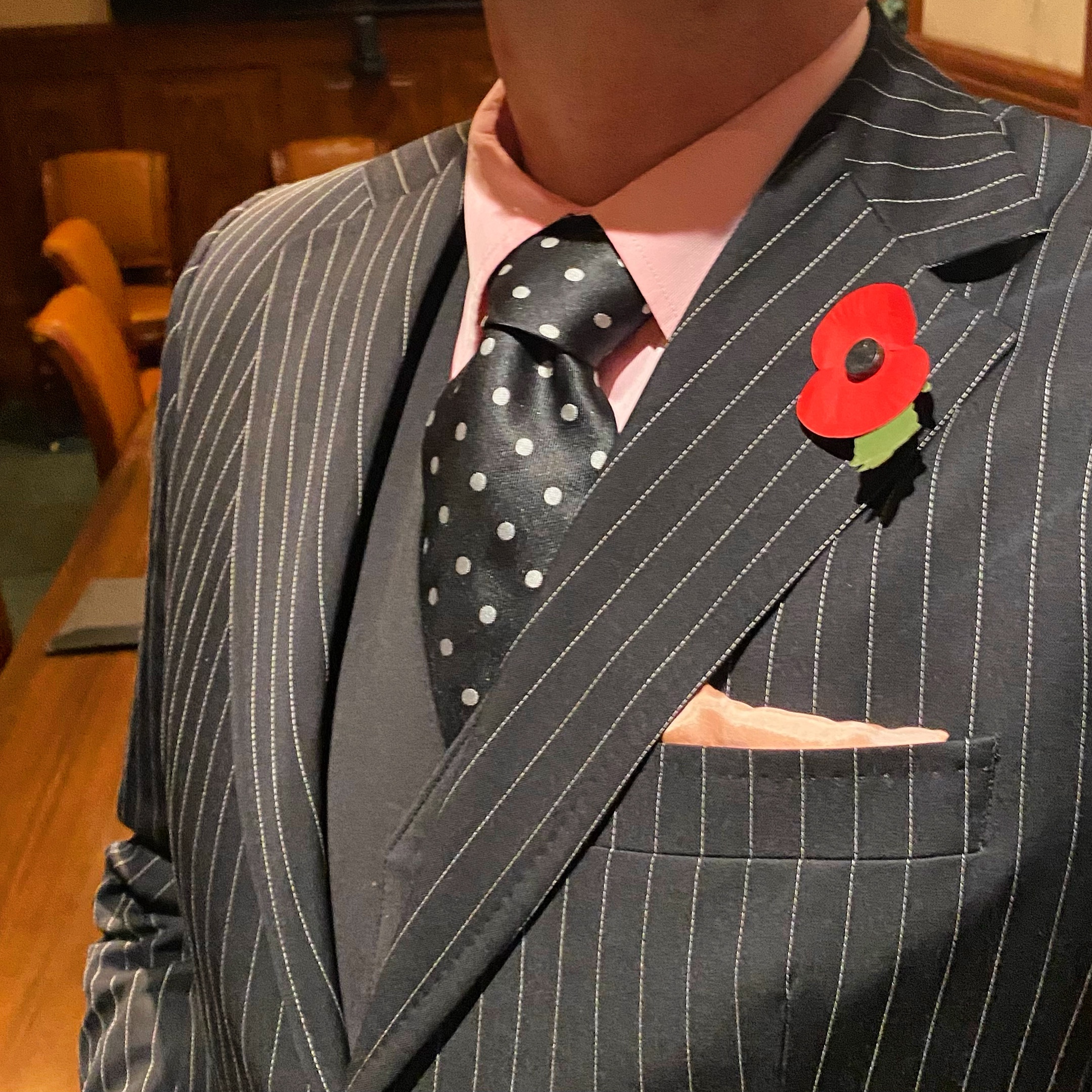 Man wearing a pinstriped suit with a pink shirt and polka dot tie, adorned with a red poppy brooch.