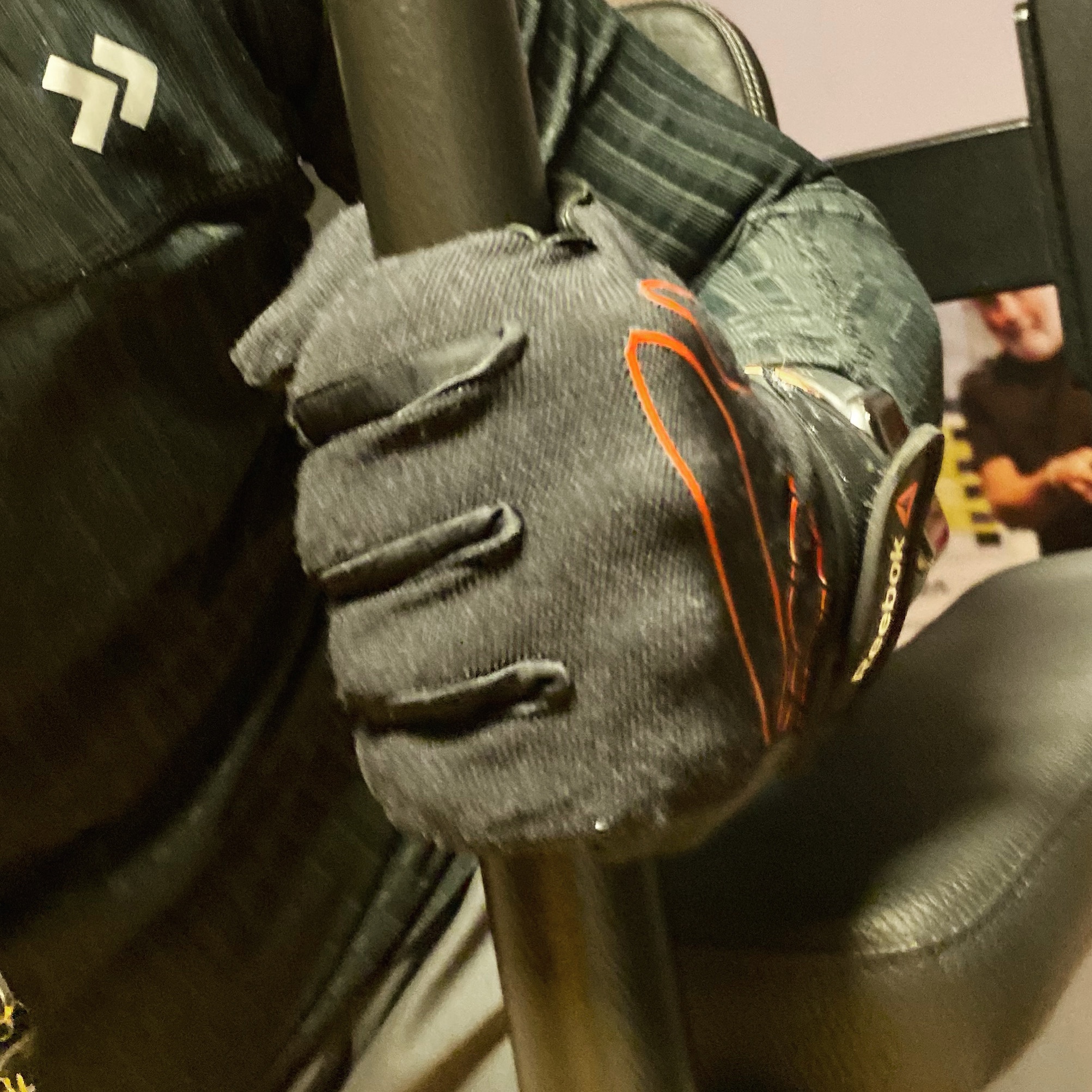 Person wearing a black glove holding a pole with a textured grip in a gym setting.