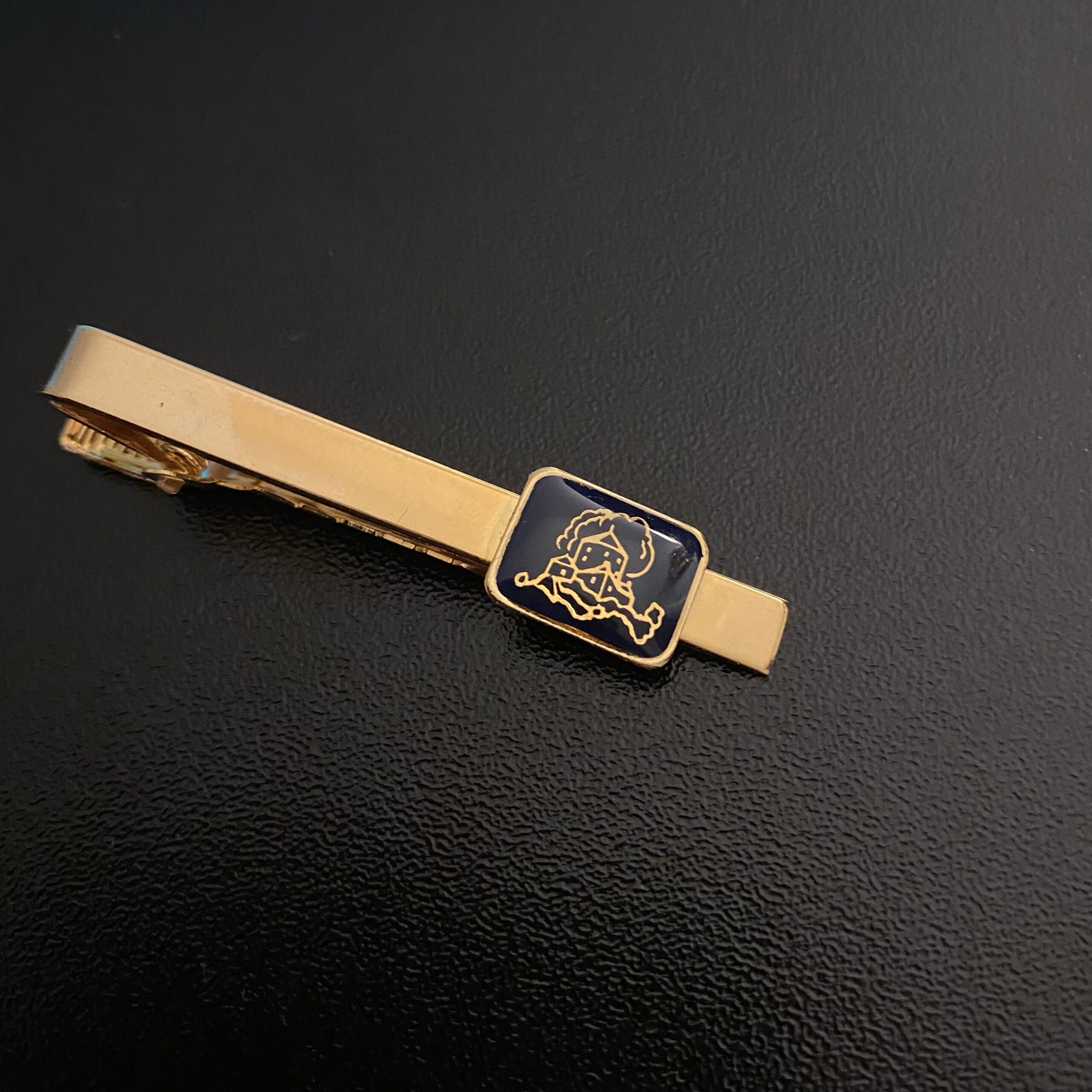 Gold tie clip with a black enamel inlay featuring a detailed design of a banker's emblem.