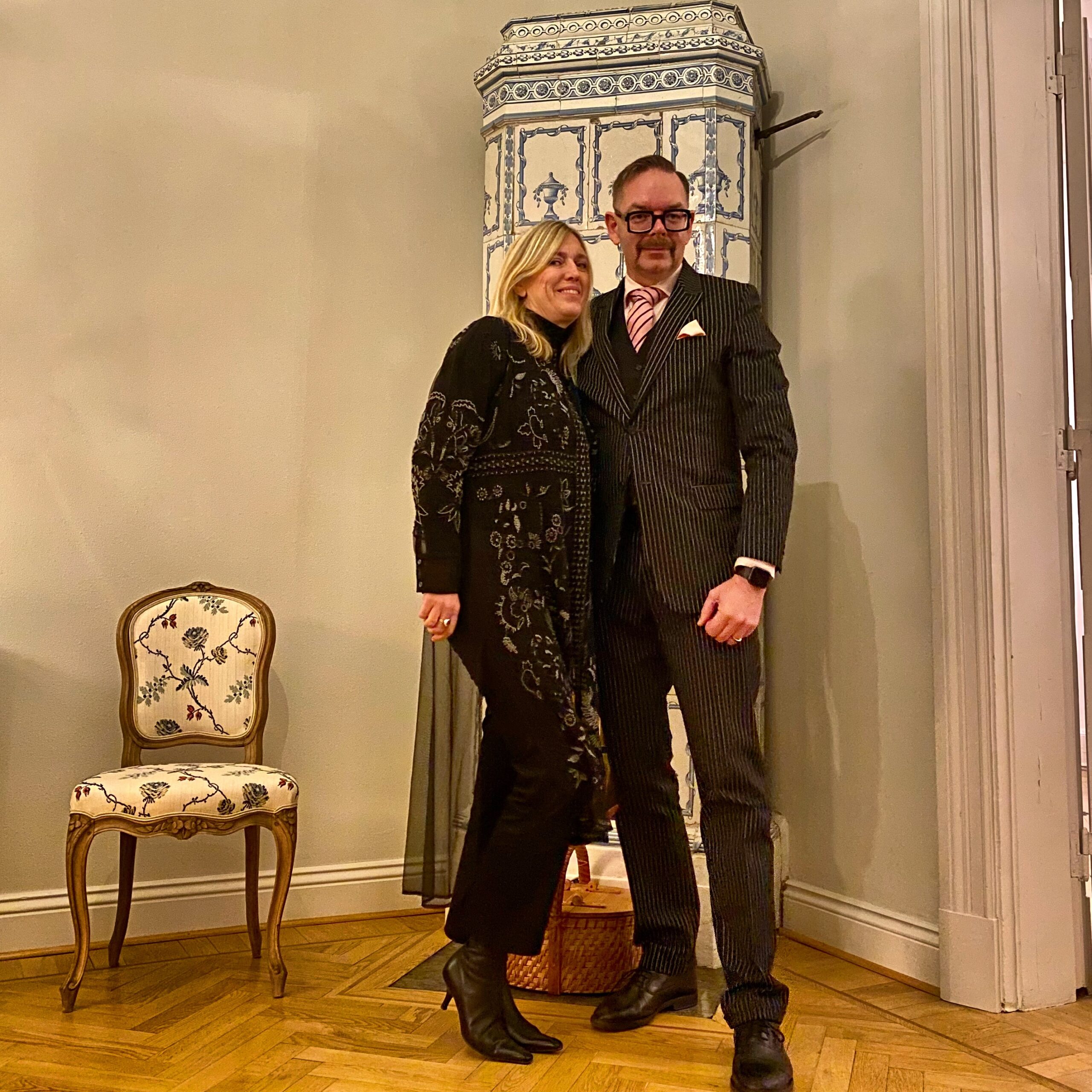 A man in a striped suit and a woman in a black embellished dress posing together in an indoor setting.