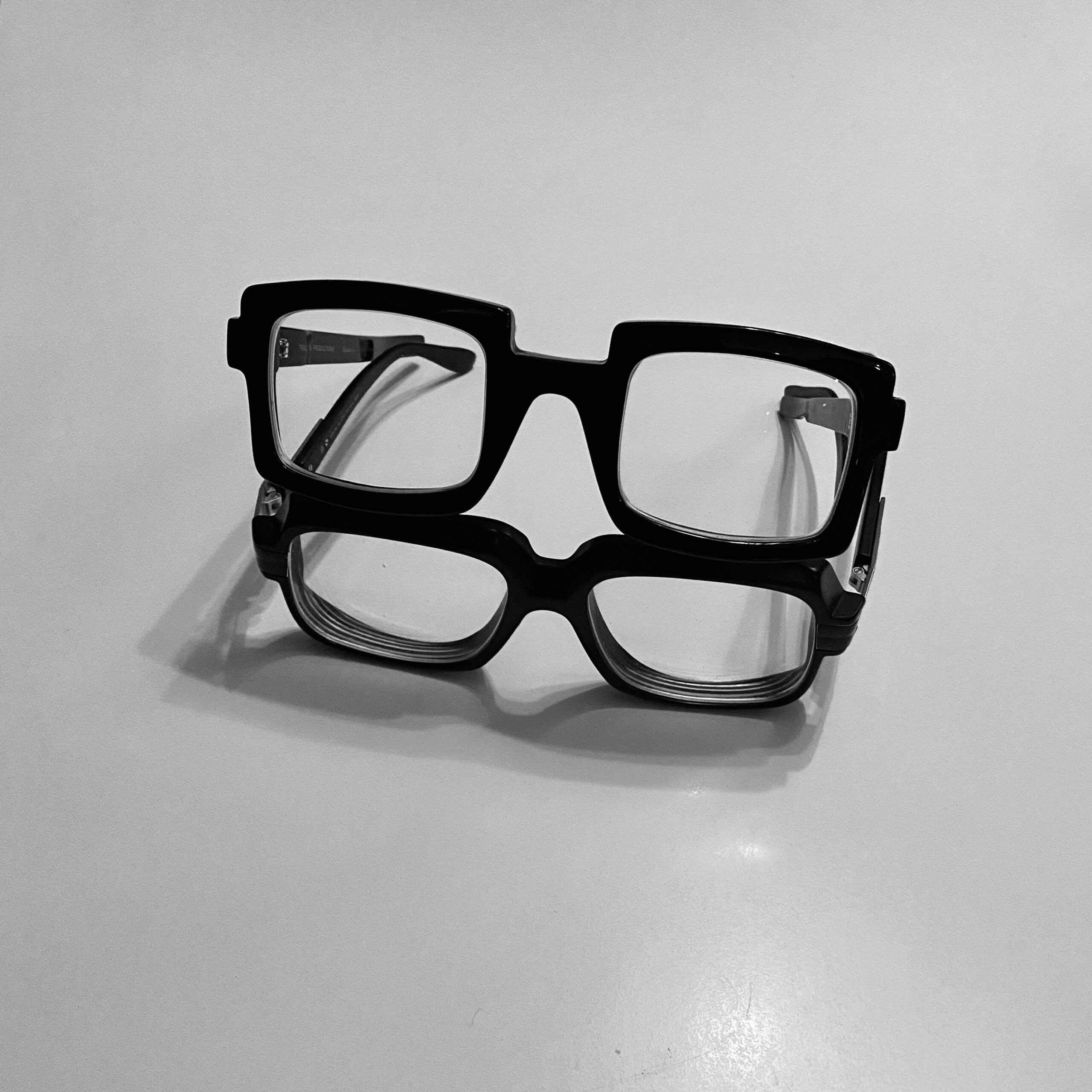 Two pairs of black glasses stacked on a flat surface, showcasing distinct styles and sizes.