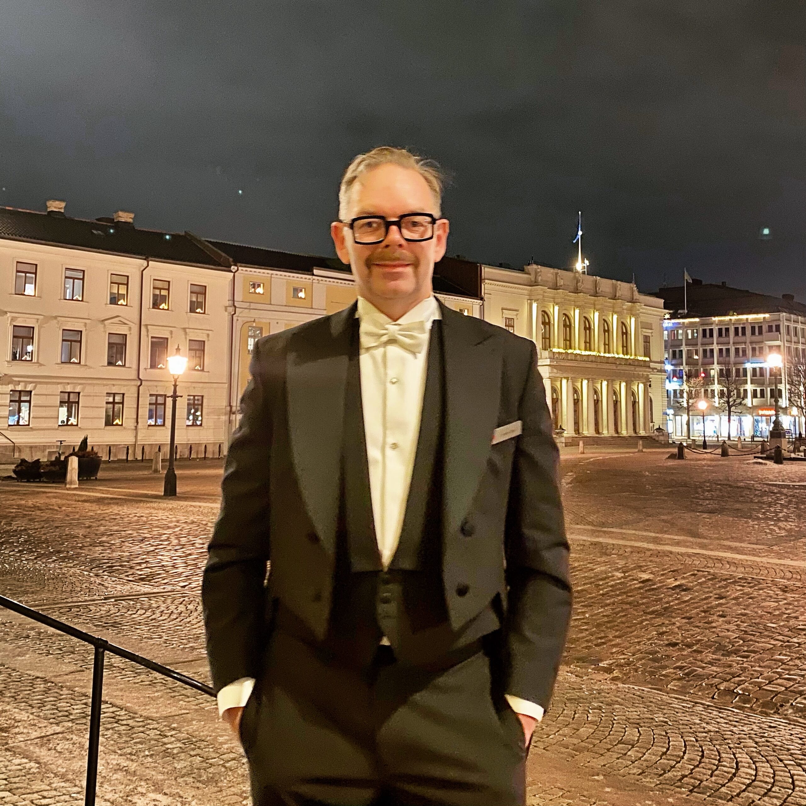 Man in formal evening attire standing outdoors at night in a city square with historic buildings.