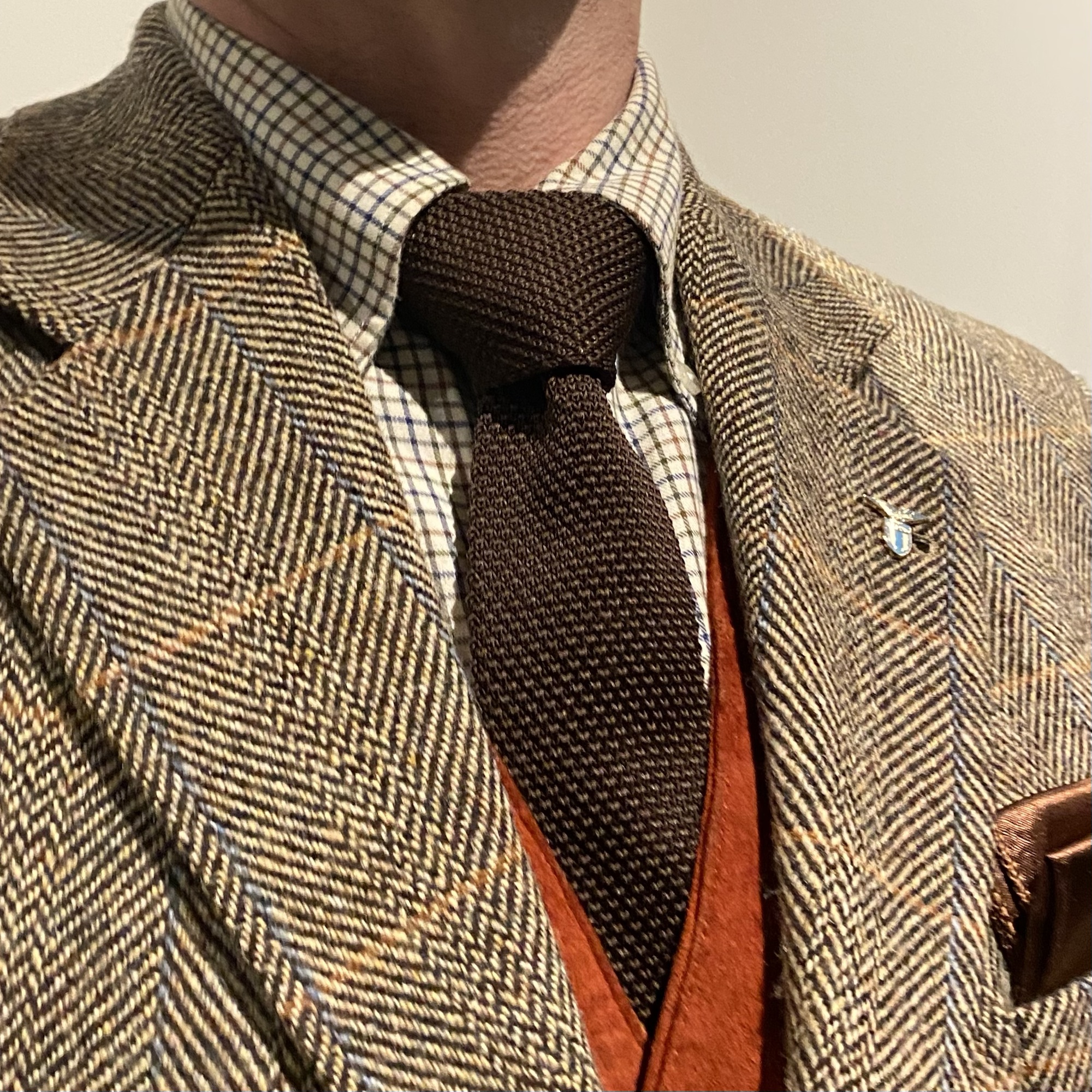 Man wearing a patterned brown tweed jacket with a checked shirt and a knitted tie, showcasing a classic style.