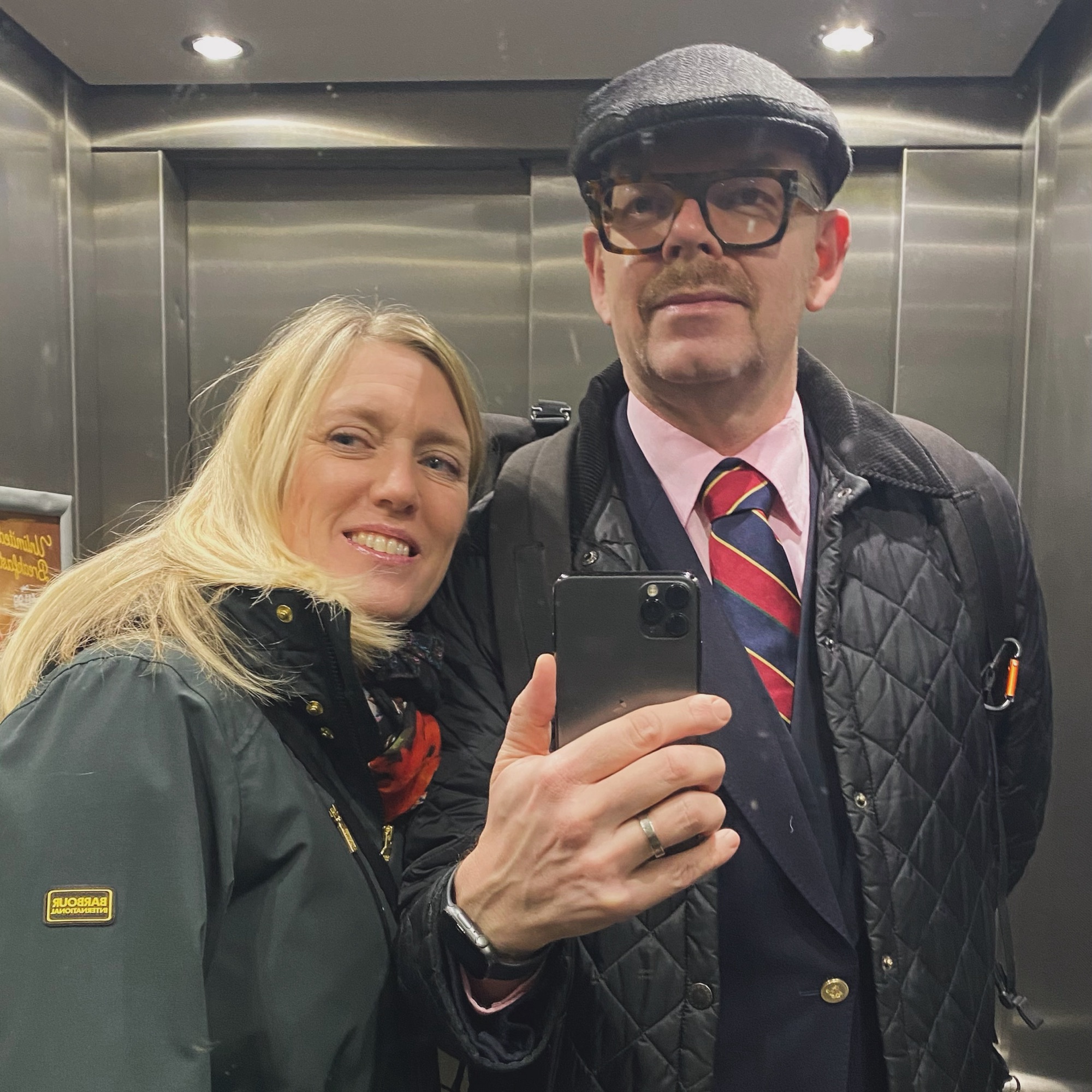 A man in a cap and glasses alongside a woman with long blonde hair, both smiling in a mirror selfie in a lift.