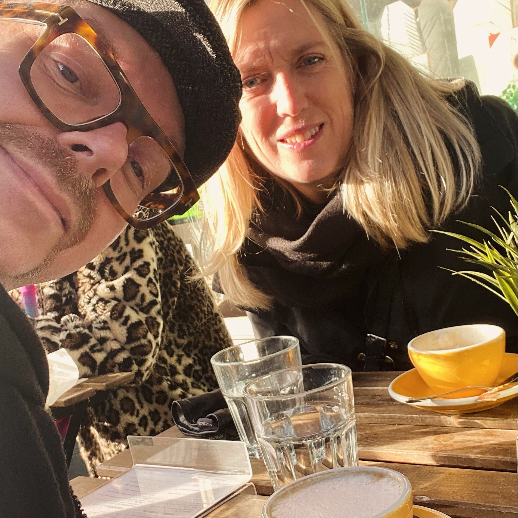 Two individuals seated at a table with drinks, one wearing glasses and a hat, the other with long hair and a scarf.