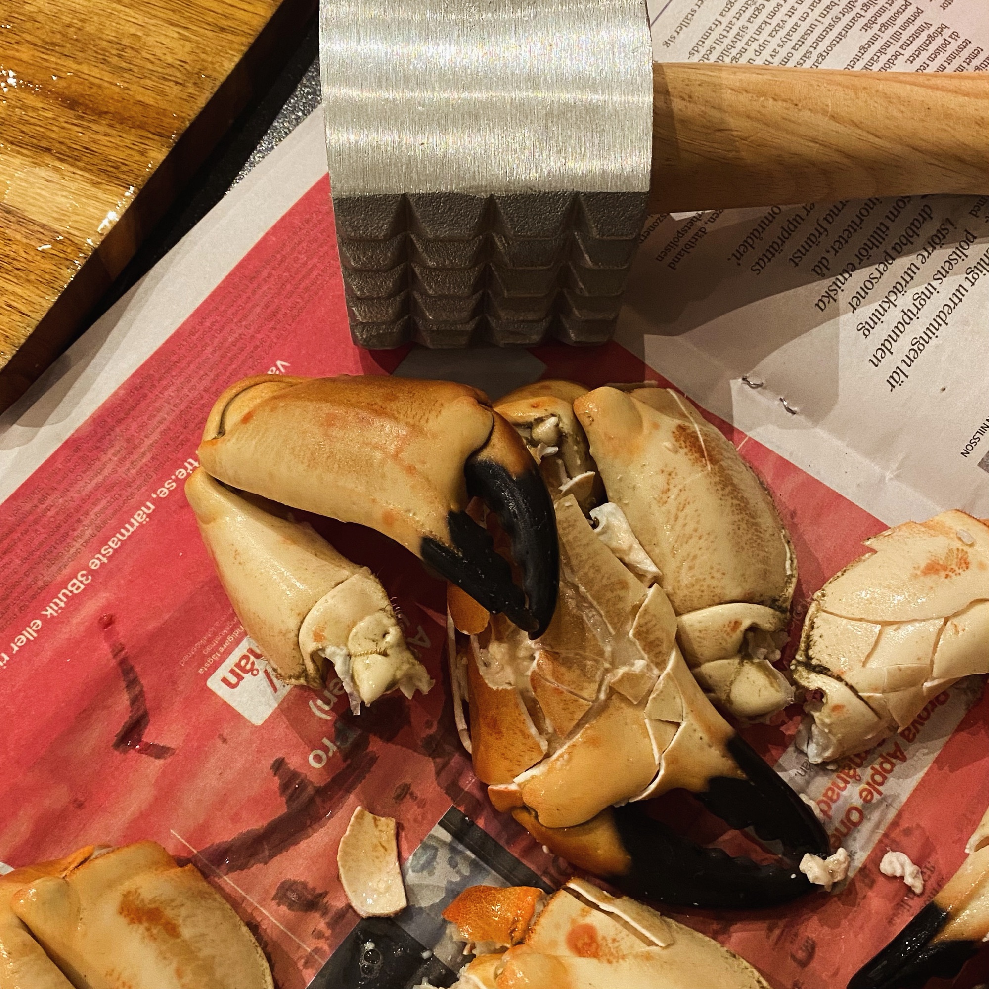 Crab claws arranged on a newspaper with a metal crab mallet resting nearby.