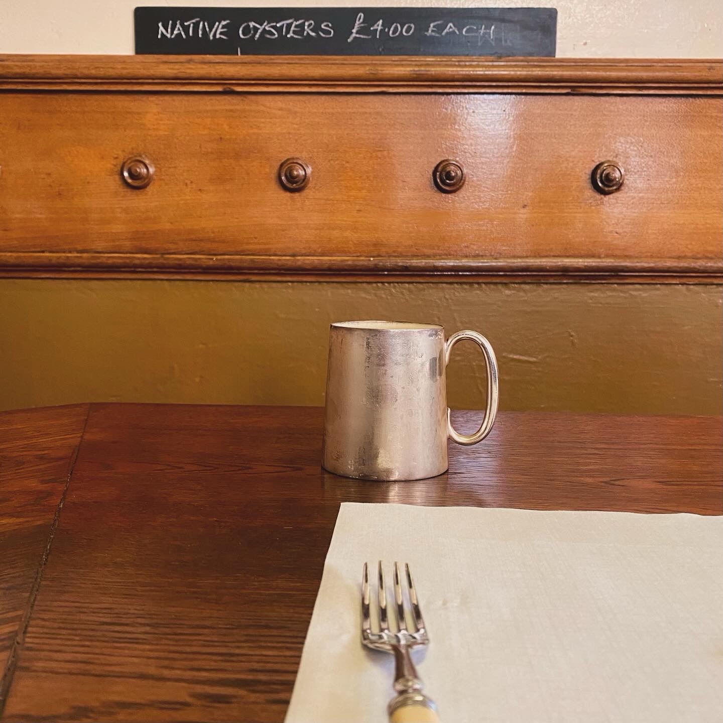 A silver mug and a fork placed on a wooden table with a chalkboard menu in the background listing native oysters.