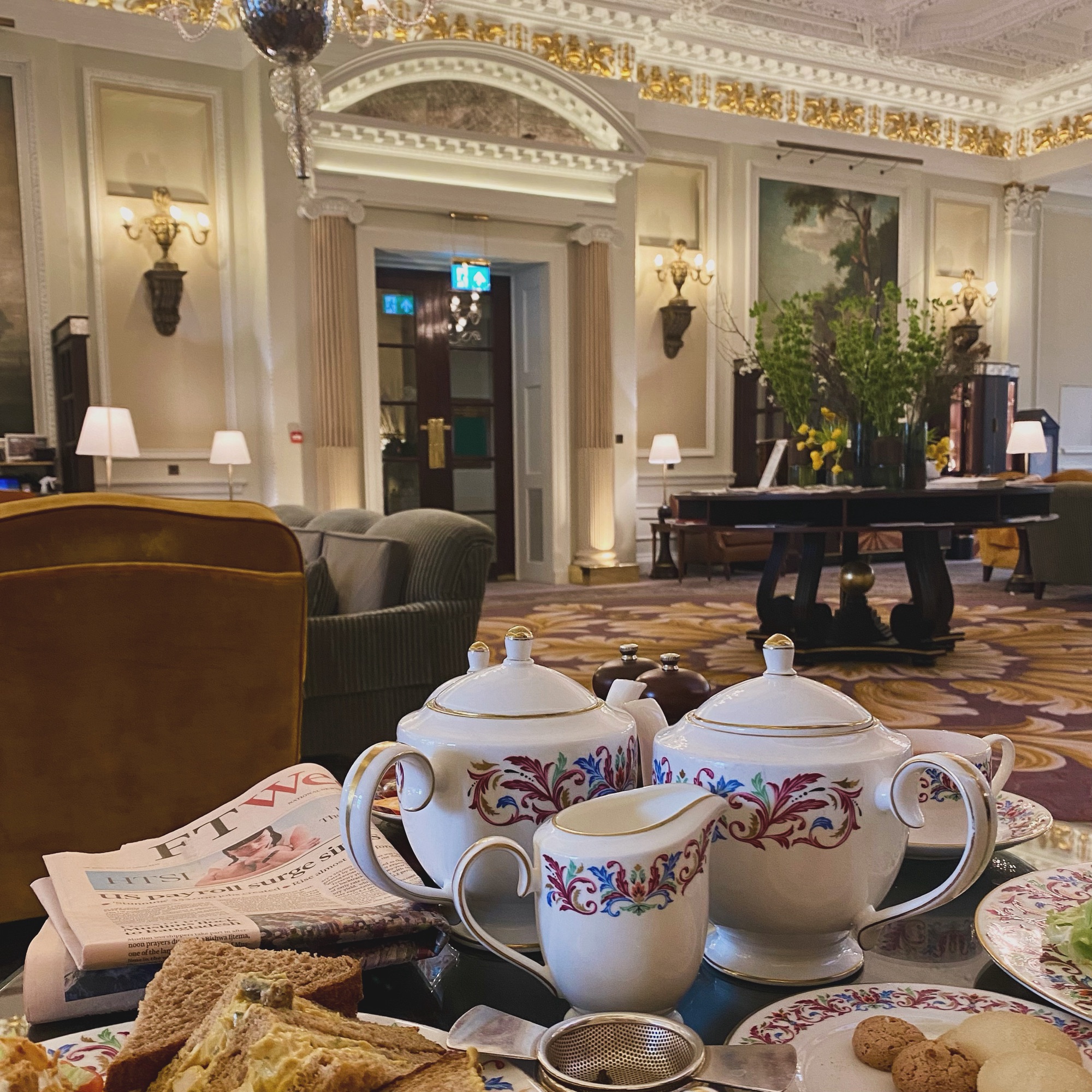 Elegant tea set with floral design, accompanied by sandwiches and biscuits on a table in a sophisticated indoor setting.