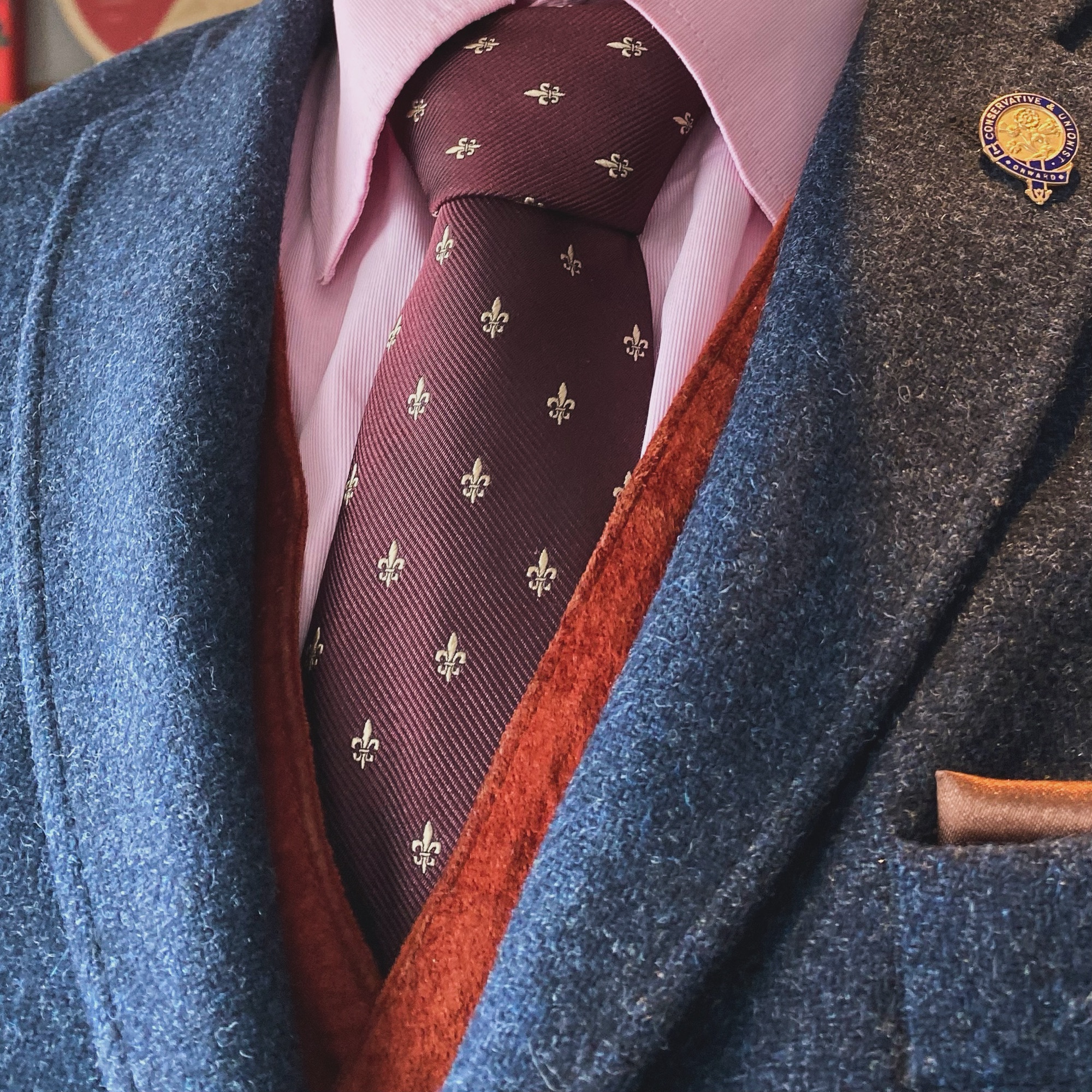Close-up of a man wearing a navy blue blazer, maroon tie with fleur-de-lis pattern, and a pink shirt.