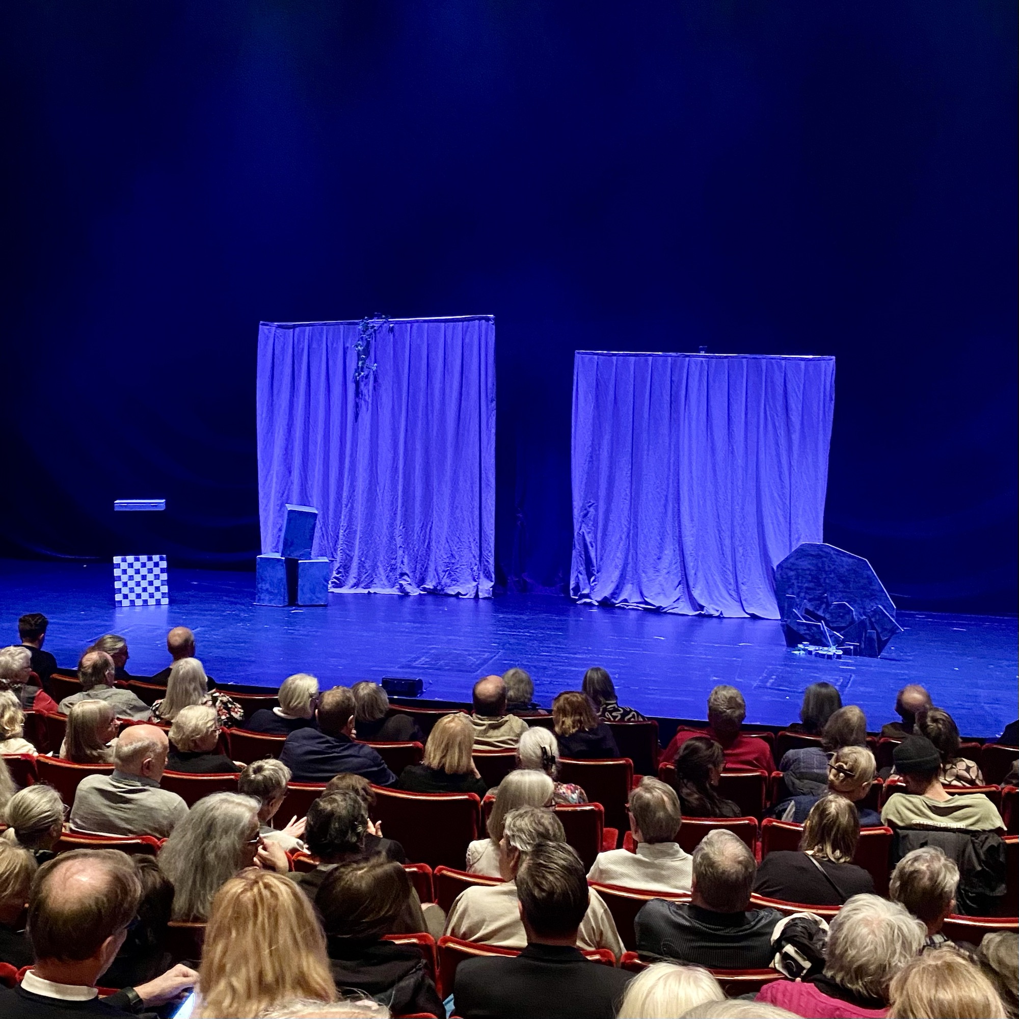 Theatre stage set with blue curtains, a chair, and a rock in front of an audience.