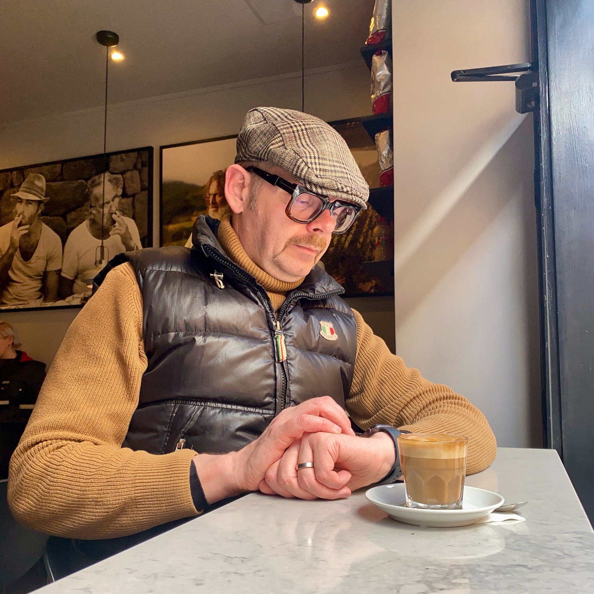 Man wearing a flat cap and glasses sitting at a table with a coffee cup in a café setting.