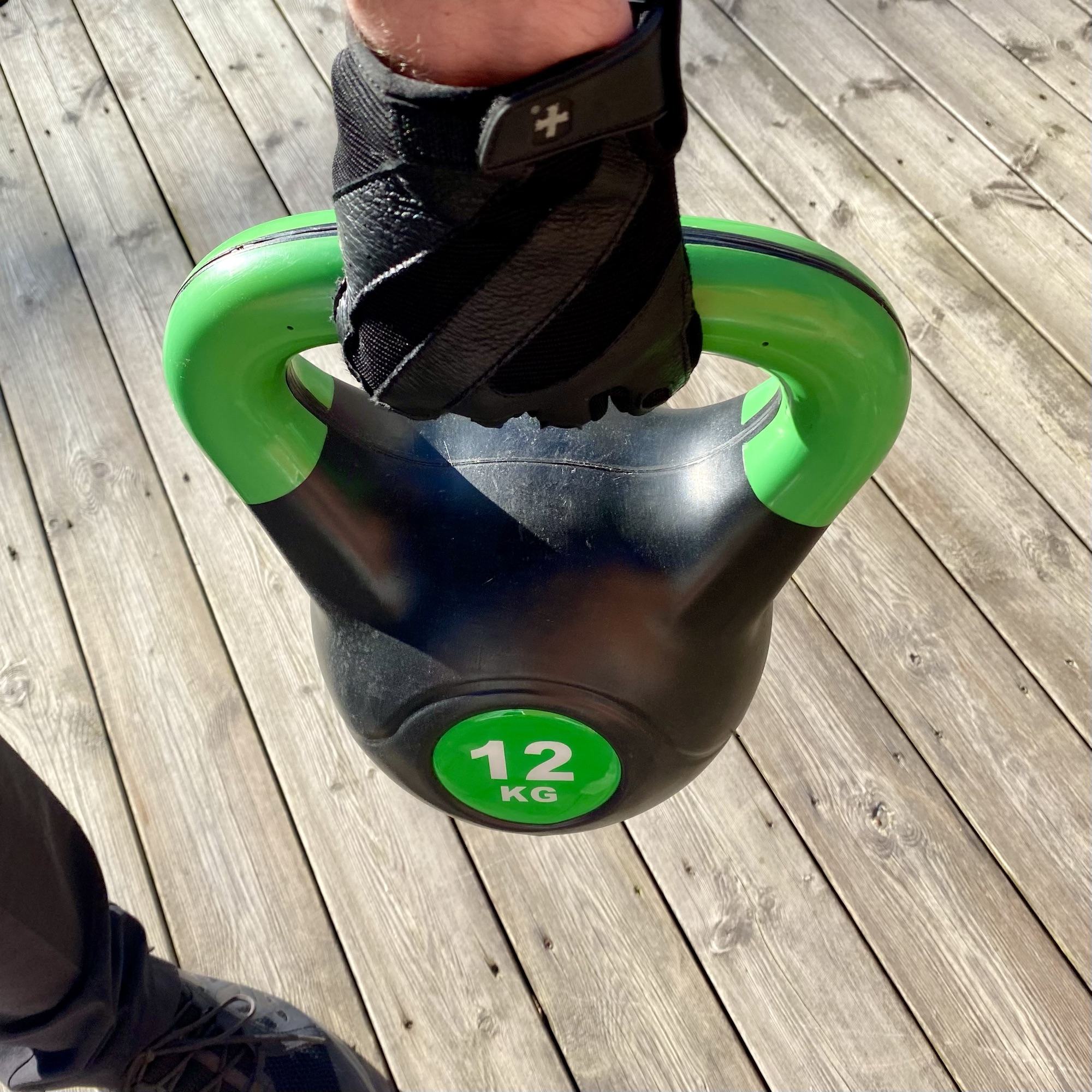A person holding a 12 kg black kettlebell with green accents on a wooden surface.