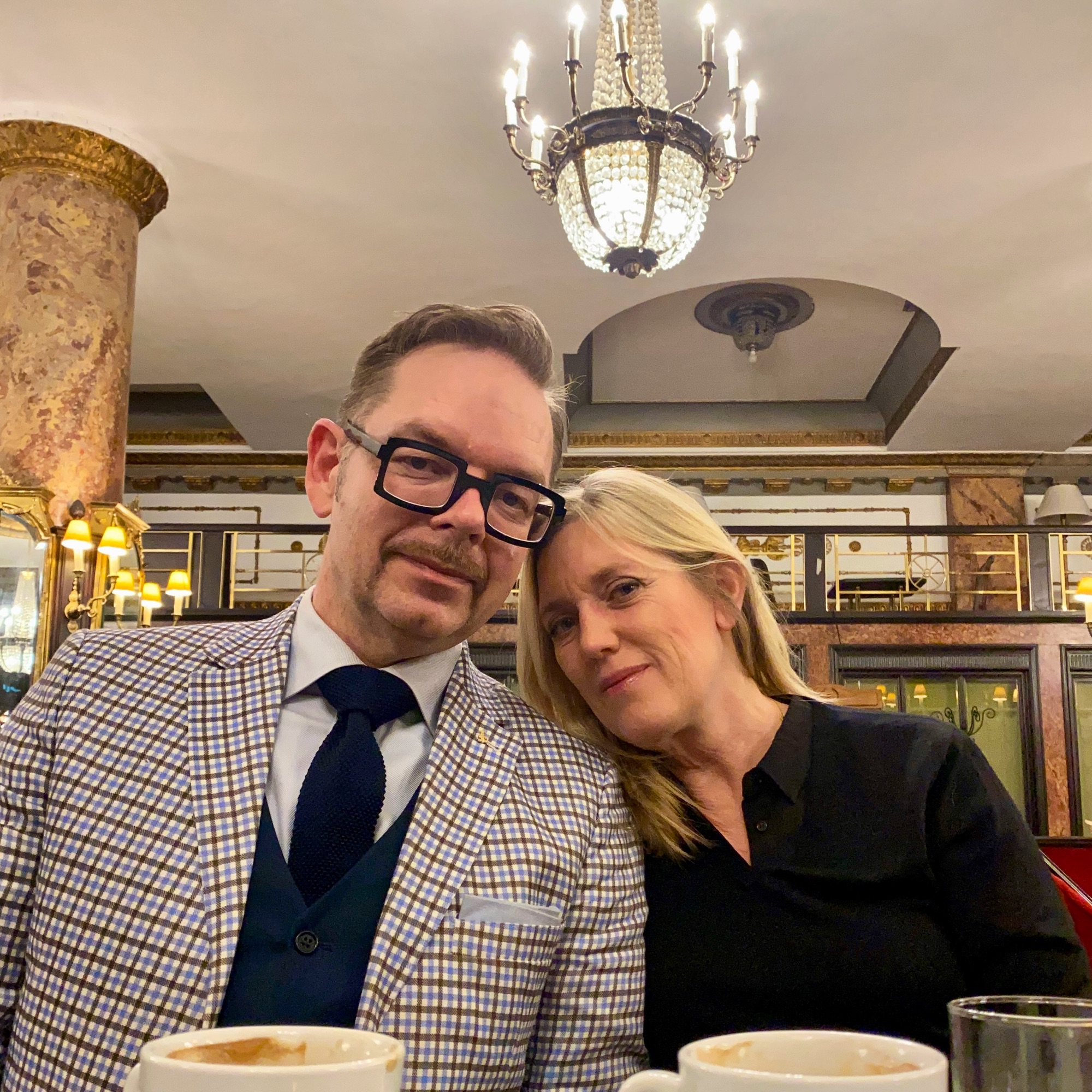 Man in a checked suit and woman in a black top seated together in a café with coffee cups.
